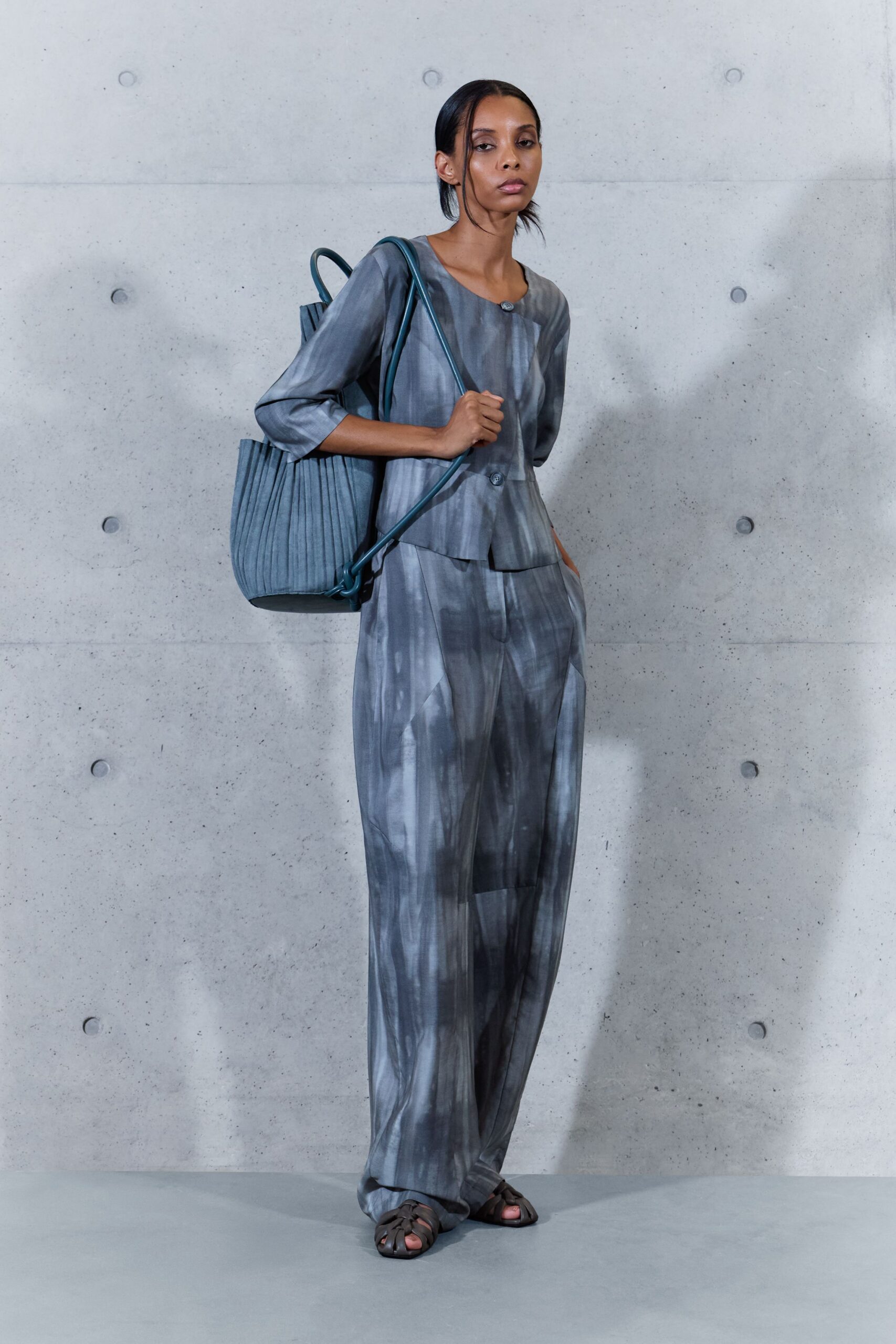 Smoky grey top with a round neck and two mottled buttons. Matching wide trousers in blurred brushstroke print, pooling over the sandal. Slate pleated tote with a long tubular shoulder strap. Dark leather fisher sandals. Crossing a museum courtyard just after the paving dries from rain.