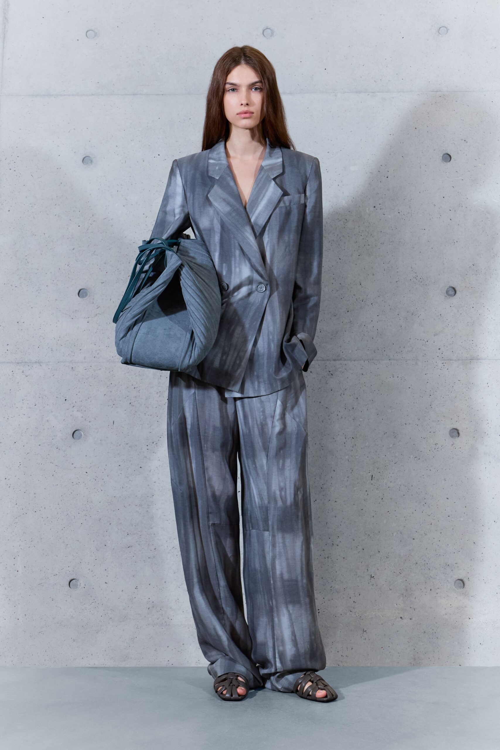Double-breasted blazer in smoky grey satin, washed with blurred vertical streaks. Slouchy slate shoulder bag, sides pinched into deep pleats. Wide trousers in the same streaked satin, pooling over the instep. Brown cage sandals with rounded straps. Walking across a corporate plaza just after the rain dried.