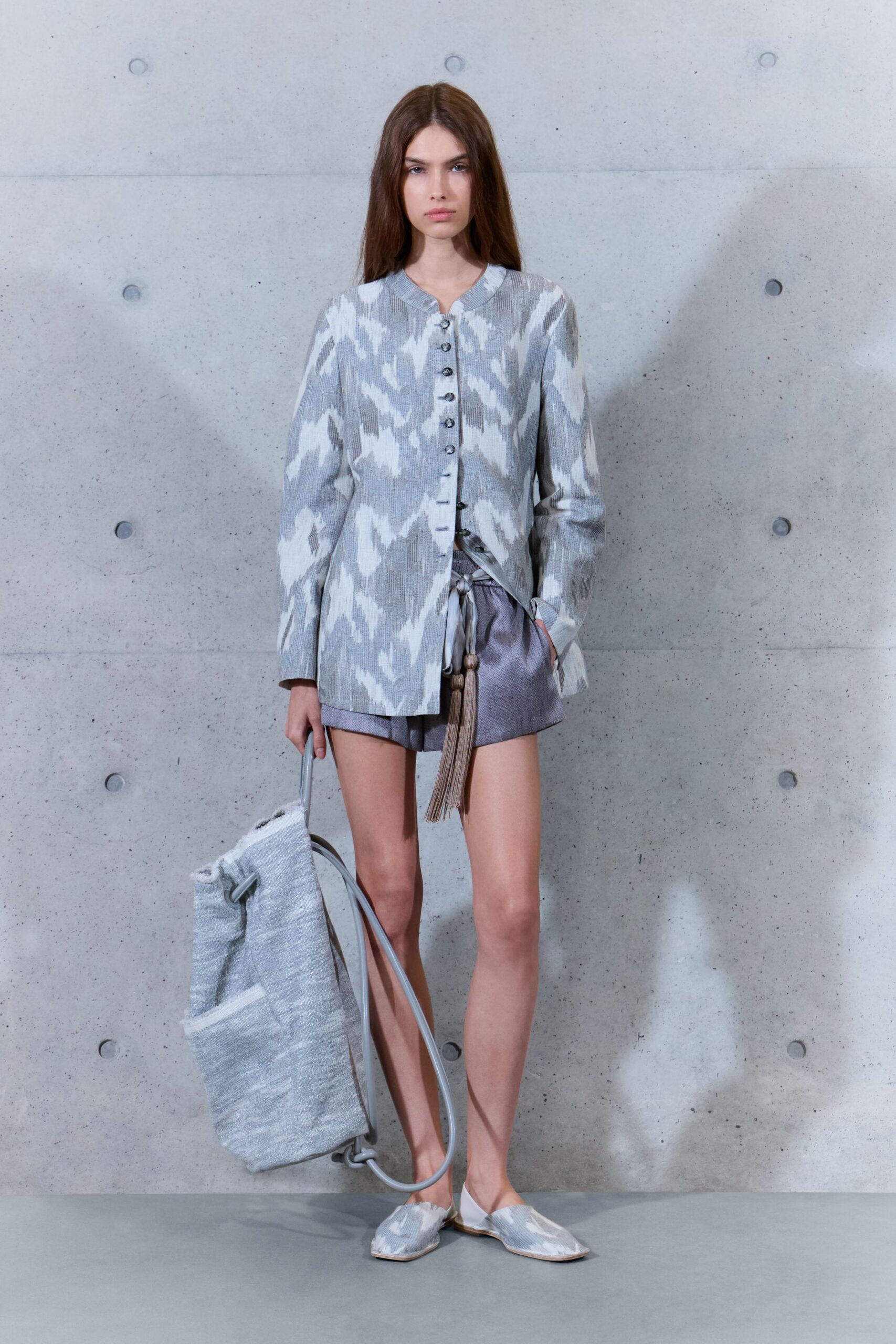 Grey-and-white ikat jacket in slub weave, tiny ring buttons running off-center. Silvery boxer shorts with a drawstring tie ending in long tassels. Mottled woven tote hanging from a pale tubular frame. Flat mules in the same blurred ikat. Wheeling damp towels back from the pool before lunch.