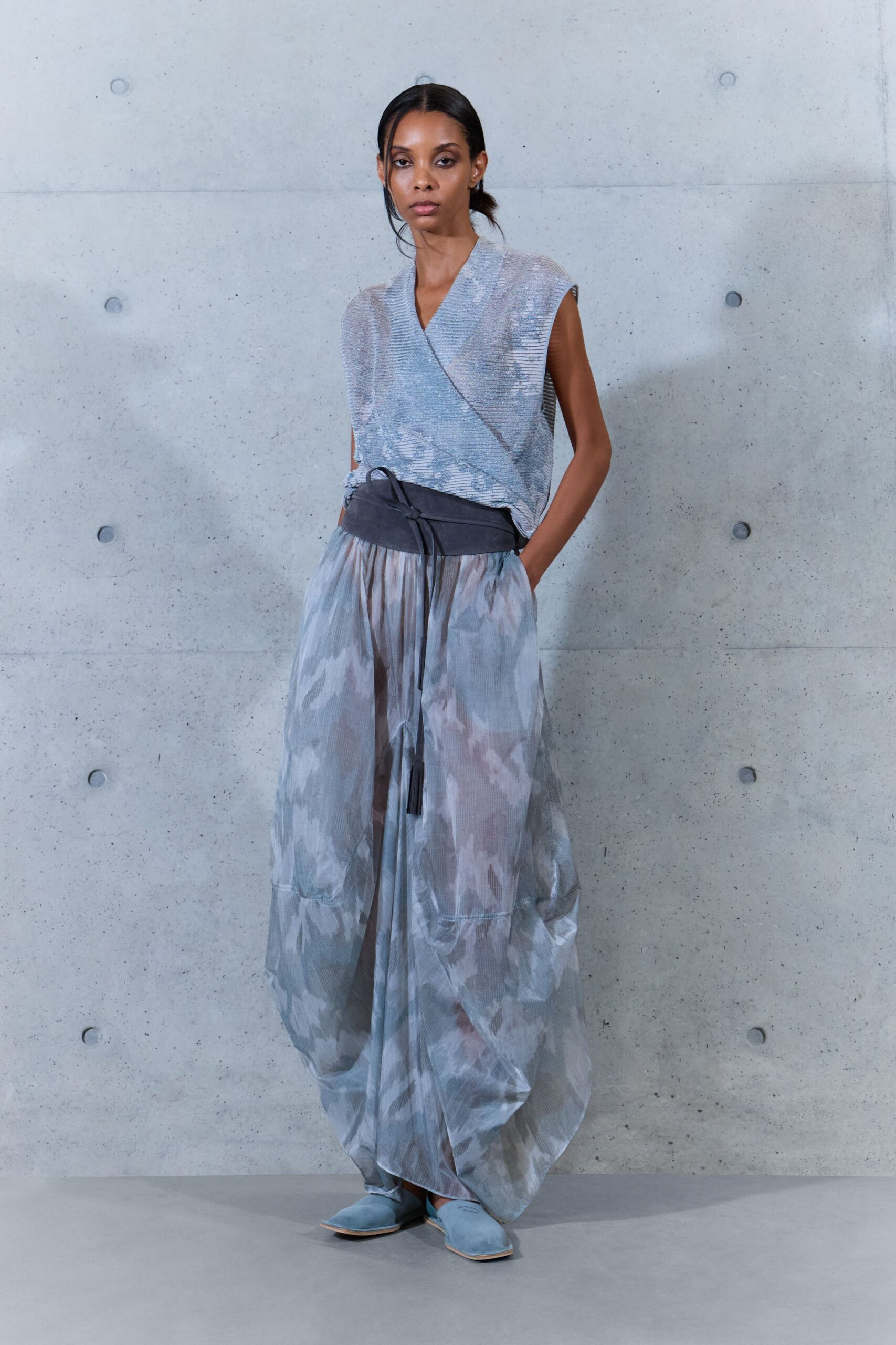 Silver-grey crinkled knit wrap top with a washed clouded shimmer. Voluminous sheer trousers in the same misted print, gathered into lantern hems. Charcoal sash belt with long tasseled cords hanging off-center. Powder-blue suede flats with squared vamps. Crossing a museum courtyard just after the paving dries from rain.