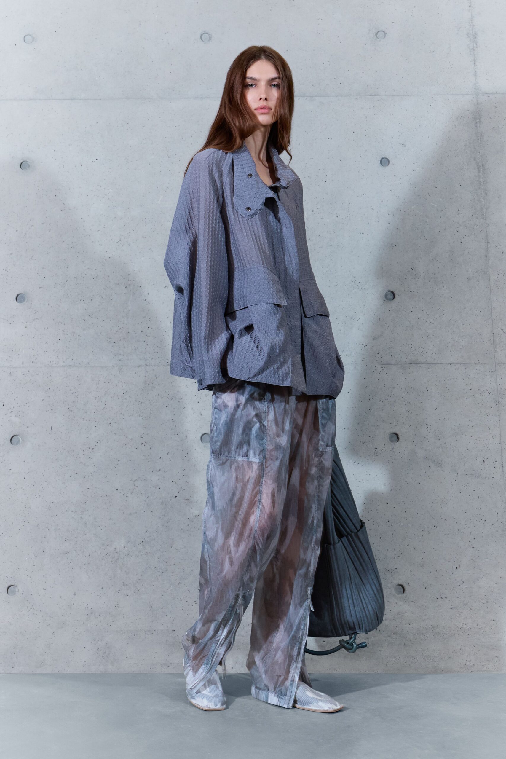 Slate crinkled jacket with a skewed snap tab and dropped sleeves. Misty sheer trousers with cloudy marbling and zip vents at the hem. Square-toe boots under gauzy printed covers. Charcoal pleated sack bag with a knotted drawcord loop. Crossing an empty concrete atrium before the building heat switches on.