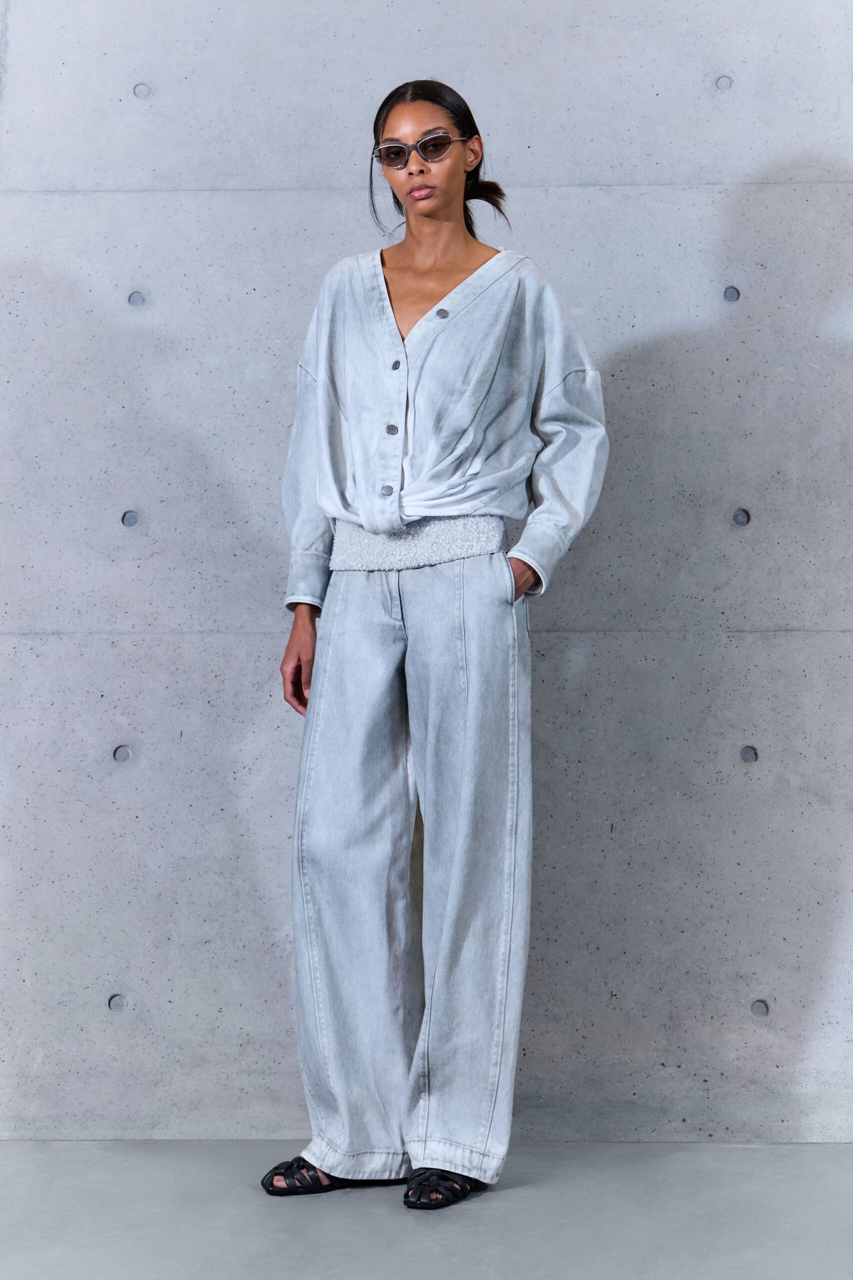 Narrow smoked lenses in thin silver frames. Bleached pale-denim blouse, dropped shoulders and a twisted placket at the waist. Matching wide-leg jeans with a nubby white waistband and long center seams. Black caged leather sandals. Walking into a concrete museum lobby before the heat has lifted.