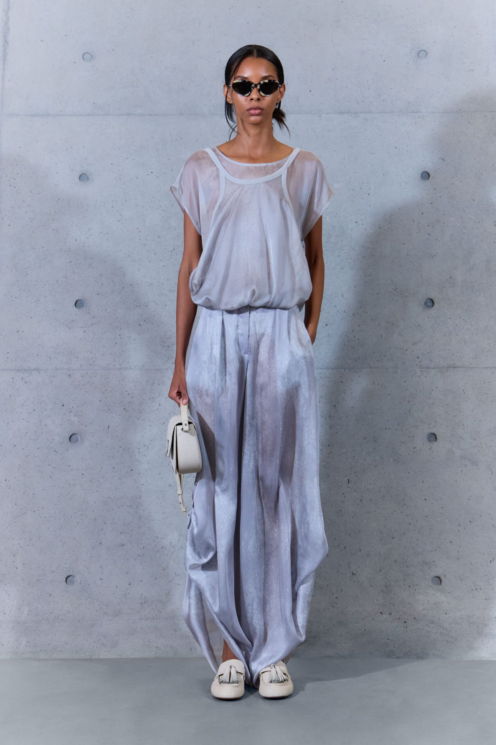 Black oval sunglasses with narrow silver arms. Sheer dove-grey top, bound seams tracing a dropped oval neckline. Pale liquid-satin trousers, full through the leg and pooling hard. Cream pebbled-leather top-handle bag. Cream loafers with long tassels brushing the vamp. Crossing a museum courtyard just after the paving dries.