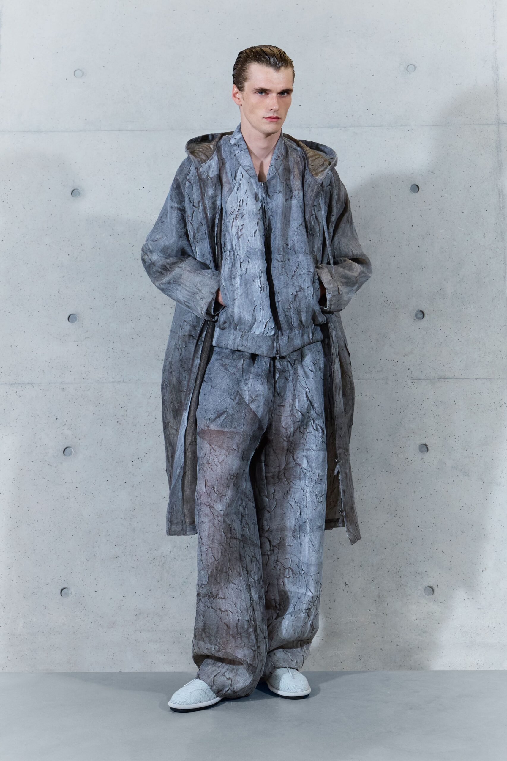 Long hooded coat in crinkled pewter-grey, open over the body. Matching crinkled zip jacket with a blunt ribbed hem. Wide trousers in the same wrinkled sheen, pooling over the foot. White quilted slippers. Walking the concrete corridor before dawn, carrying yesterday's sleep.