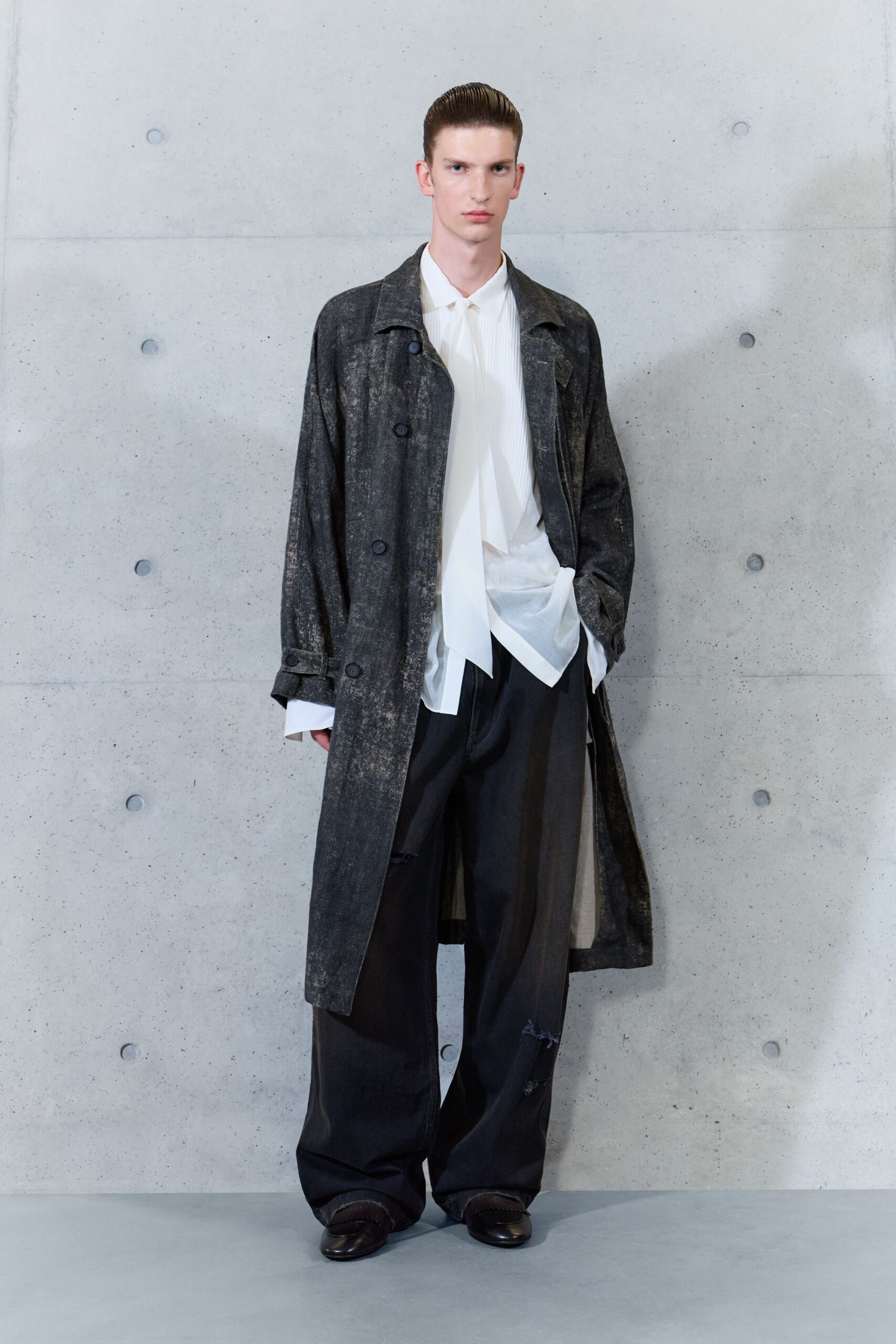 Long charcoal coat with a chalky weathered surface and loose belt tabs. Ivory pleated shirt with a long scarf panel spilling below the placket. Black wide-leg trousers, one knee torn open with loose threads. Dark leather loafers with squared apron seams. Office tower Sunday, collecting the things left in a drawer.