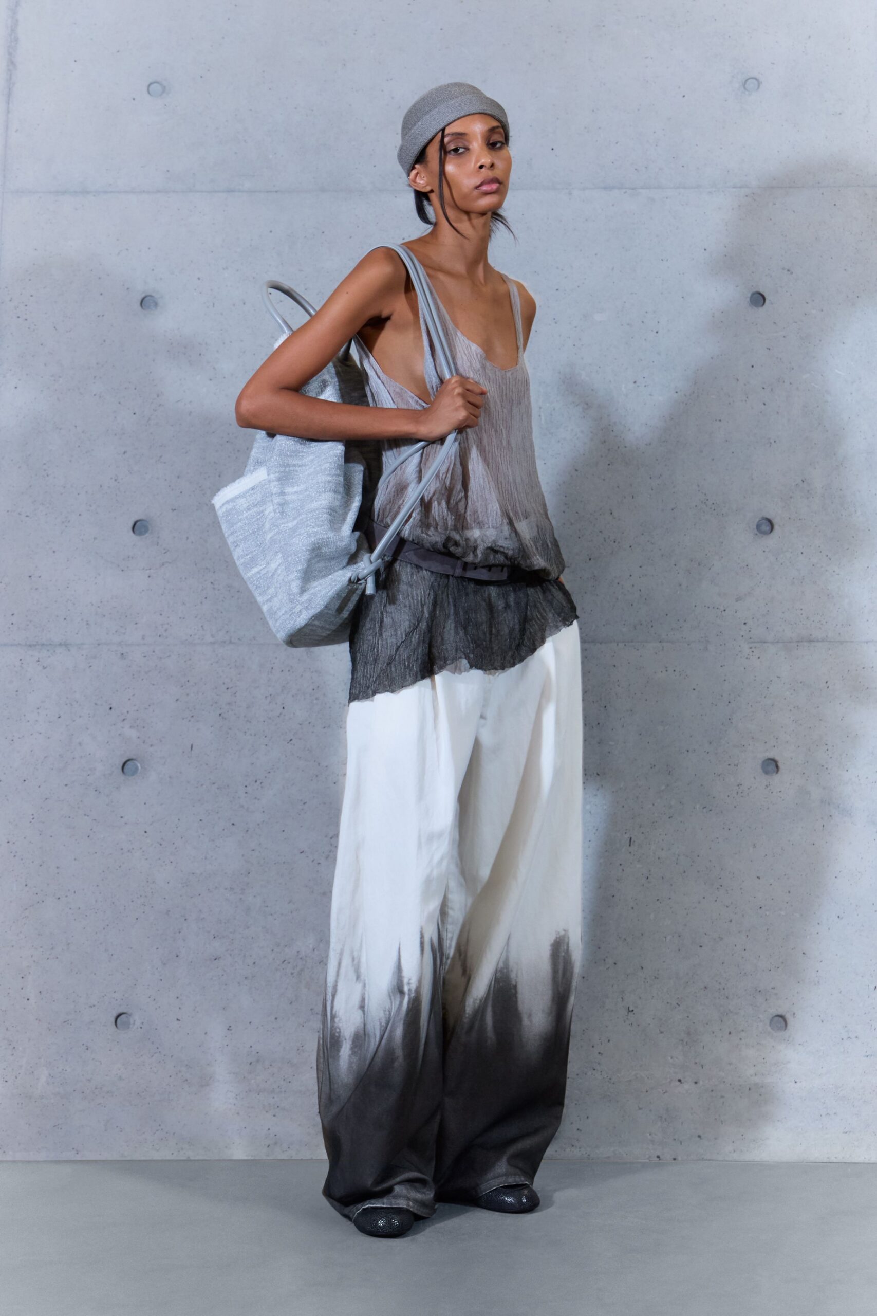 Pale grey knit beanie, rolled close above the ears. Sheer crinkled tank fading from ash to smoke, raw ruffle at the hem. Wide white trousers airbrushed to charcoal at the pooling cuffs. Large silver tote in puckered metallic weave. Dark glitter flats. Walking through a concrete courtyard before the heat burns off.