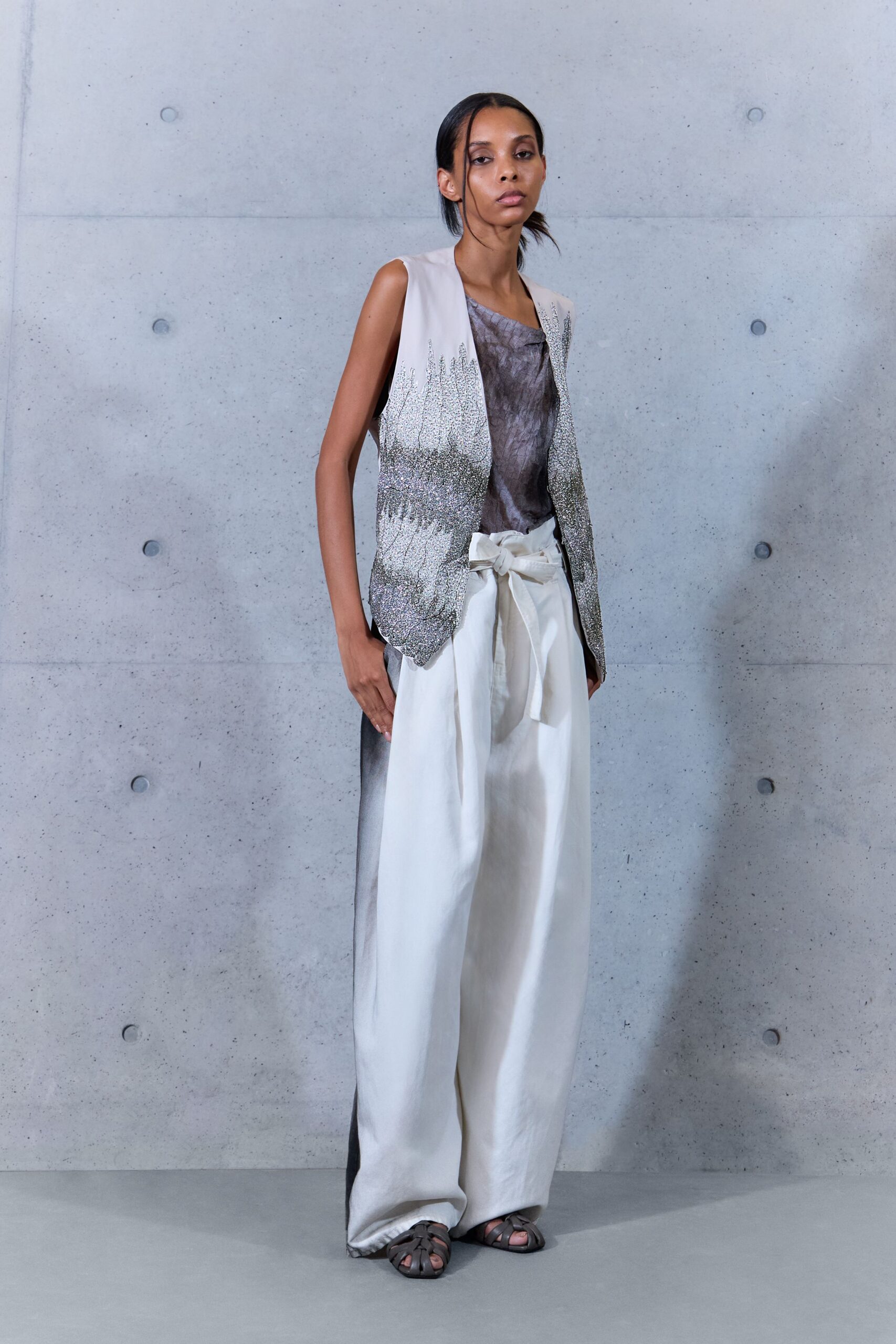 Long open vest fading from chalk to gunmetal beading, hem softly scalloped. Smoky crinkled shell, neckline folding slightly off one shoulder. White paperbag trousers with a self-tie bow slumped at center front. Taupe cage sandals. Museum atrium at noon, the concrete still cool underfoot.