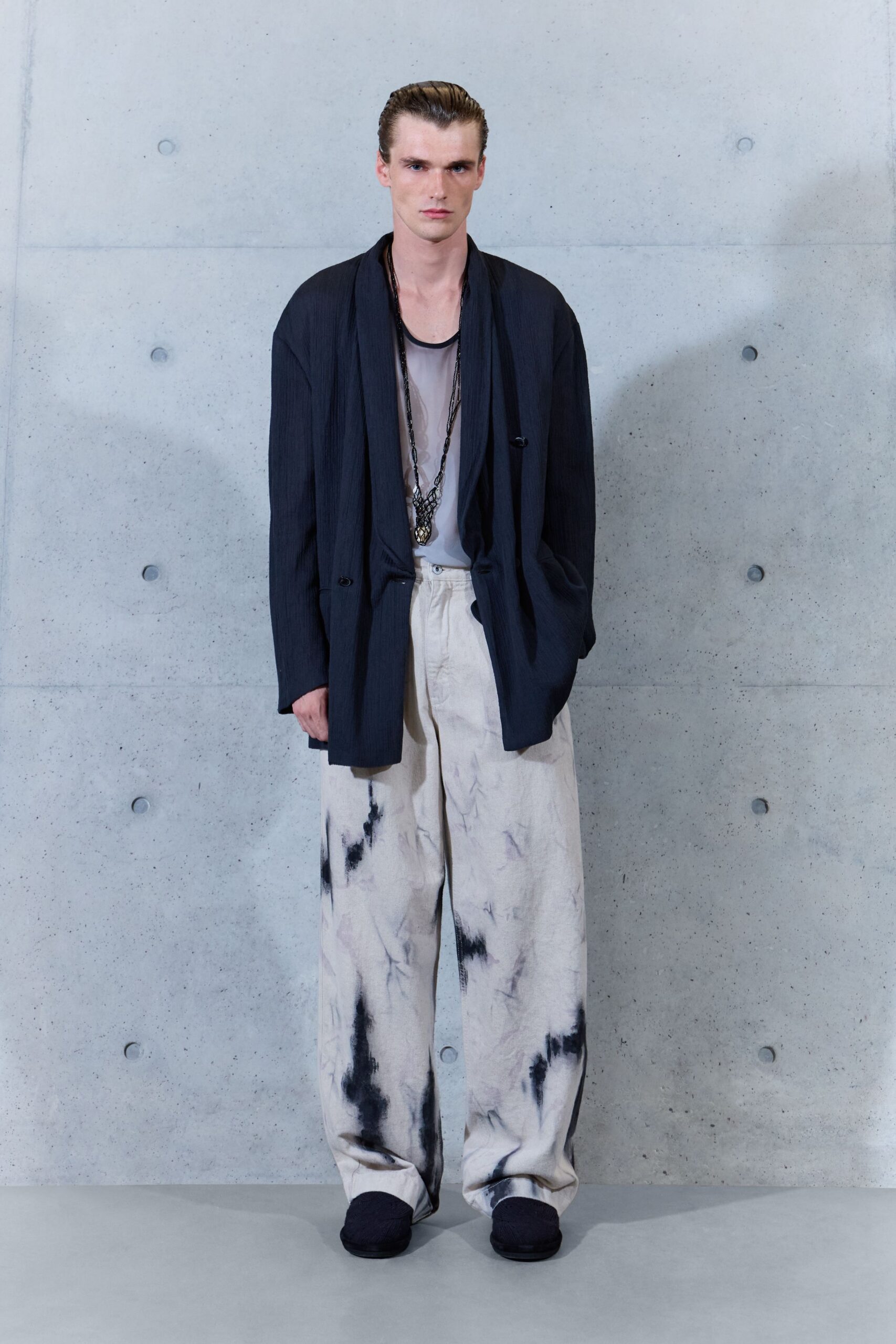 Ink pinstriped jacket with a long scarf front hanging loose. Sheer dove tank with a dark bound scoop neckline. Black netted pendant grazing the sternum. Chalk wide-leg trousers washed with smoky black blooms. Dark quilted slip-ons. For crossing a museum courtyard just after the rain stopped.