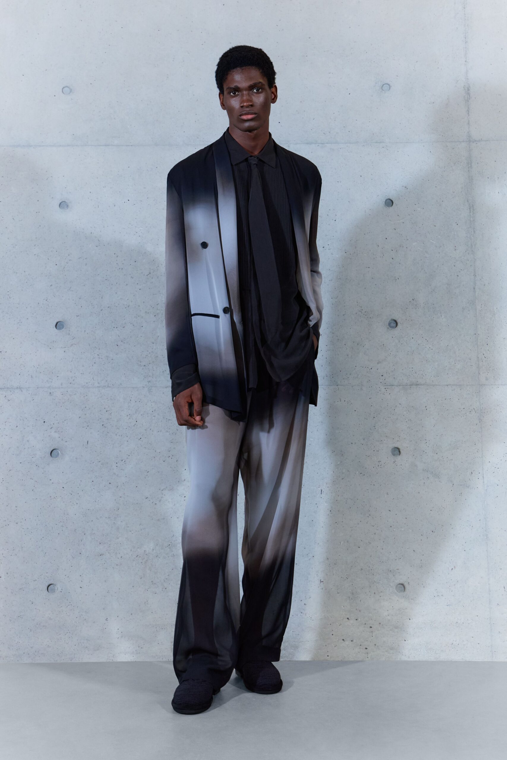 Long double-breasted blazer airbrushed from black into foggy grey. Black plissé shirt with a narrow tie panel hanging long. Wide trousers in the same smoky fade, pooling over the shoe. Black slip-ons with a ribbed knit surface. Waiting by the lift after midnight, office tower still humming.