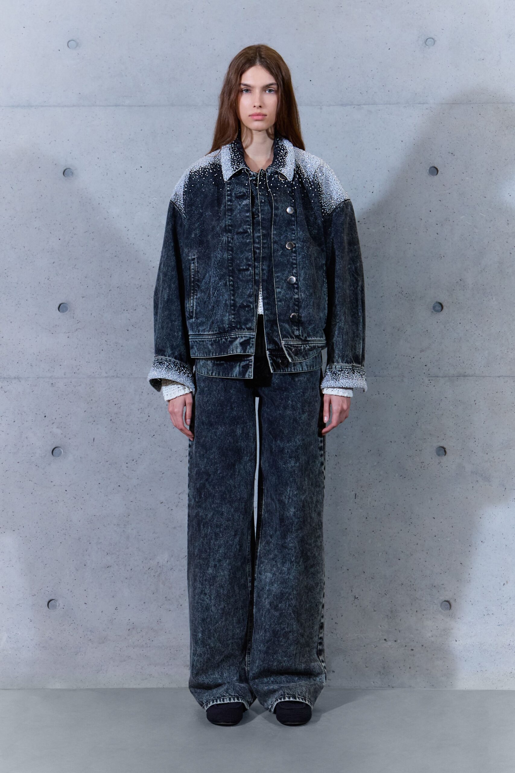 Acid-wash black denim jacket dusted with silver beading across the yoke. White open-knit cuff peeking past the turned sleeves. Wide acid-wash jeans falling straight to a pooled hem. Black square-toe shoes, nearly swallowed by the denim. Office plaza at dawn, concrete still giving back the night's cold.