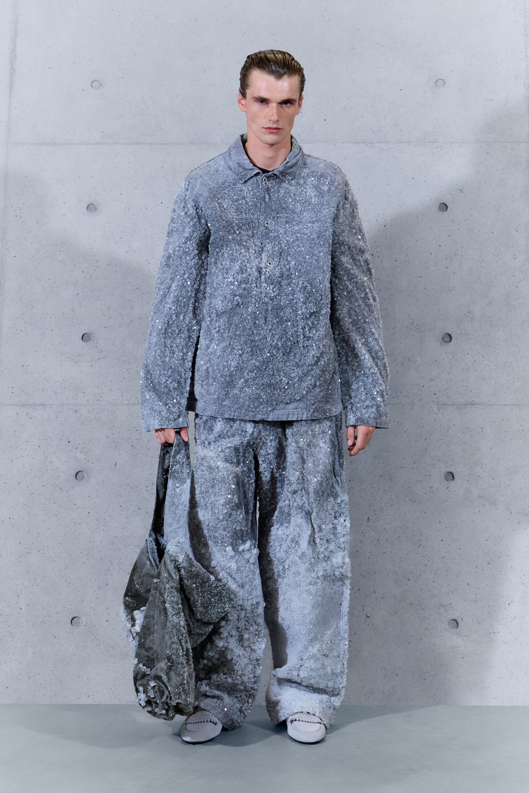 Powder-grey long-sleeve polo crusted all over with silver paillettes. Matching puddled trousers, sequins thinning to bare patches at the shin. Slouching grey tote furred with the same frosted paillette surface. Pale slip-ons with pinked apron seams. After the art school's winter gala, carrying the melted centerpieces home.
