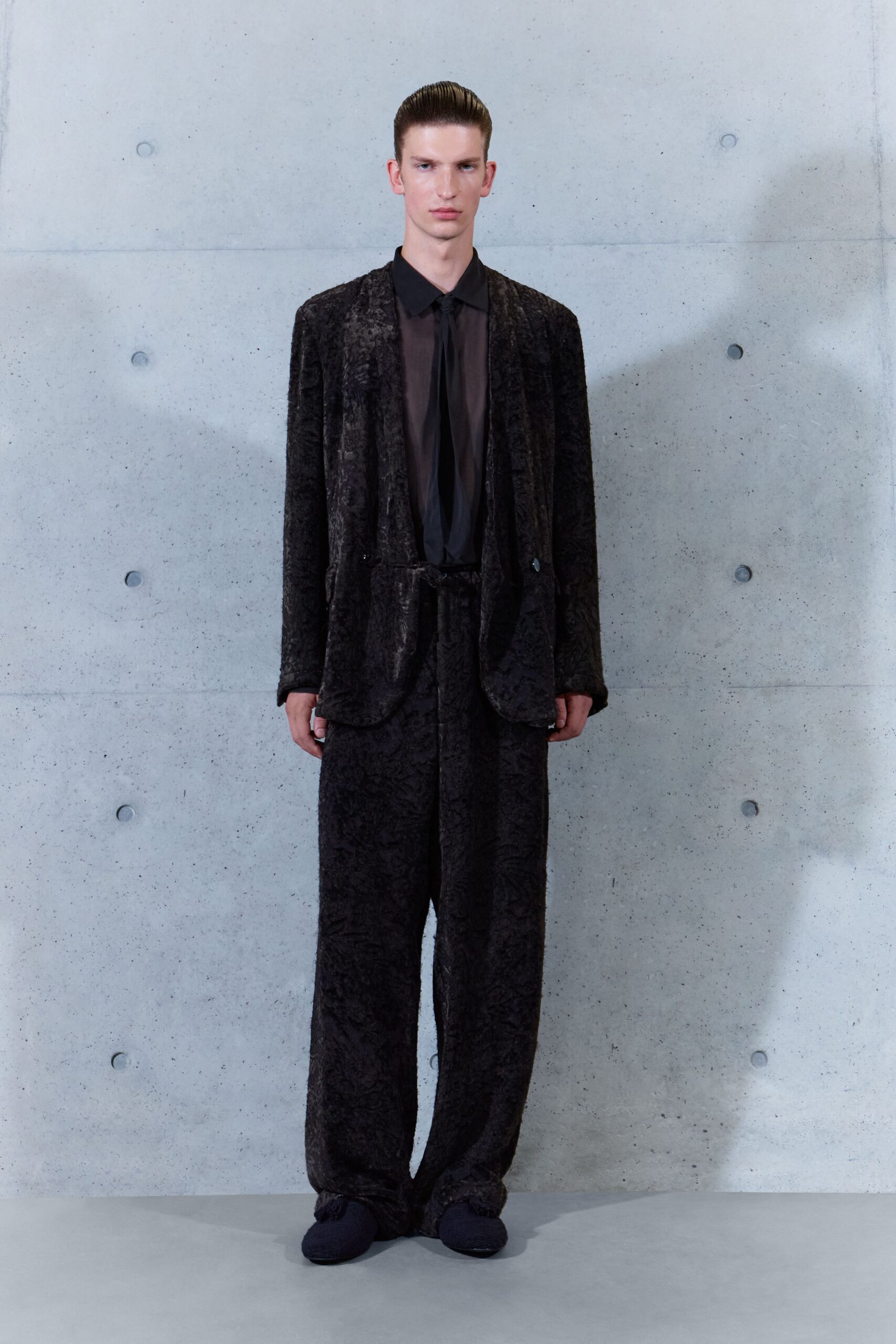 Black crushed velvet blazer with rounded patch pockets and a single button. Sheer smoke-black shirt, collar sharp above a long narrow placket. Loose matching trousers in the same crushed velvet, pooling at the hem. Dark knitted slippers. Whatever you wear downstairs when the hotel bar has just closed.