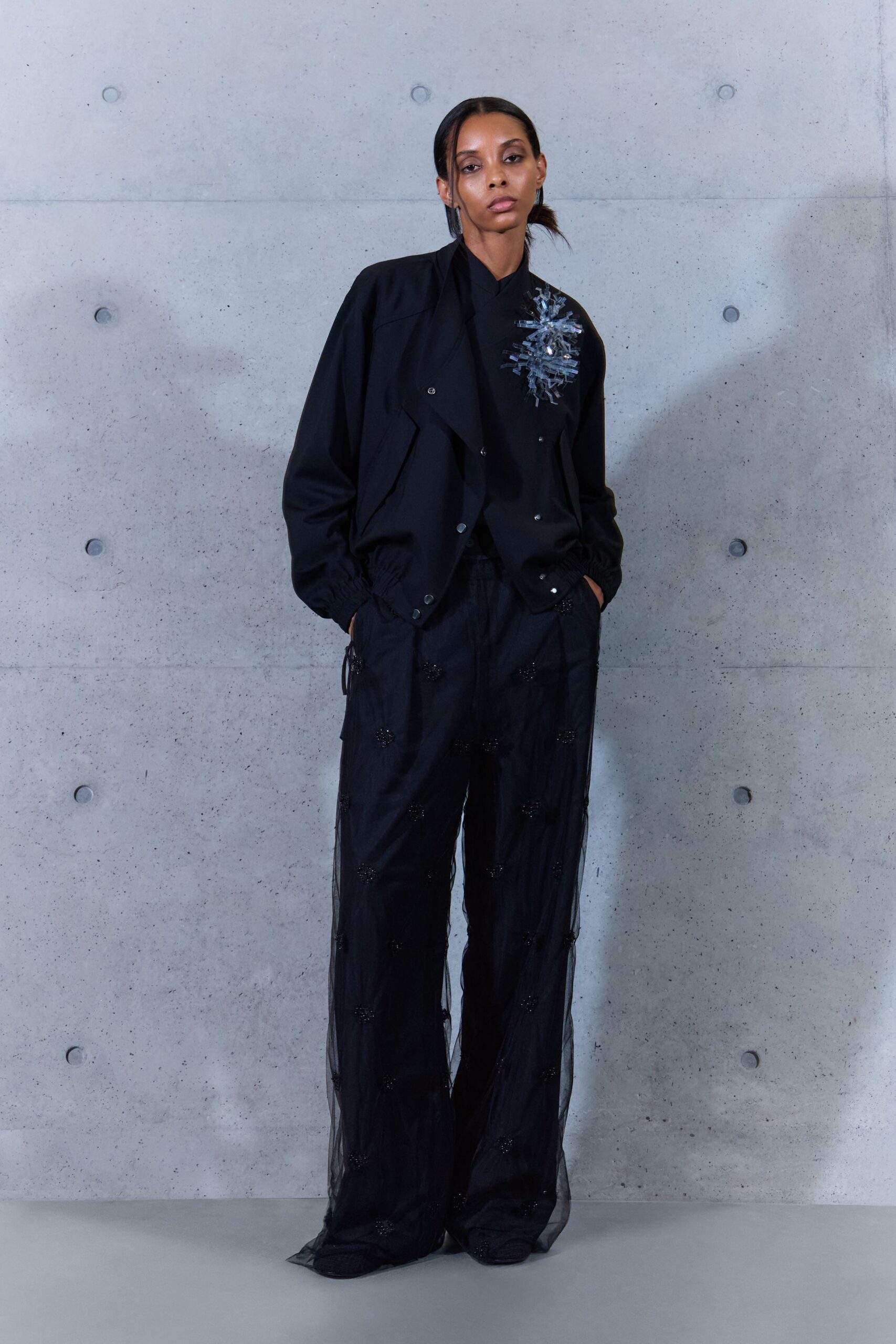 Black blouson with snapped front and elasticated hem bunching at the waist. Black high-neck top pinned with a burst of clear plastic shards. Wide black trousers veiled in sheer tulle, dotted with tiny beaded rosettes. Waiting in a concrete lobby after midnight rain, corsage catching the strip lights.