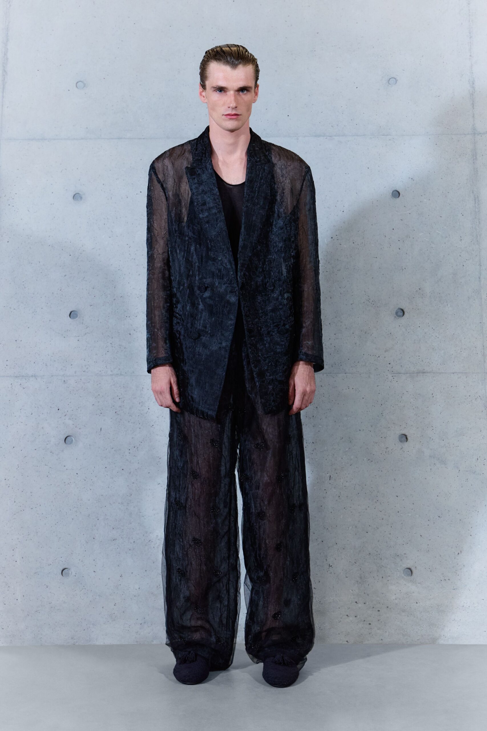 Black sheer tank, scooped close to the collarbone. Inky burnout blazer with ghosted velvet lapels and translucent sleeves. Matching gauze trousers, wide and nearly transparent with scattered dark rosettes. Black woven shoes disappearing under the hem. Crossing a hotel atrium before sunrise, still carrying last night's smoke.