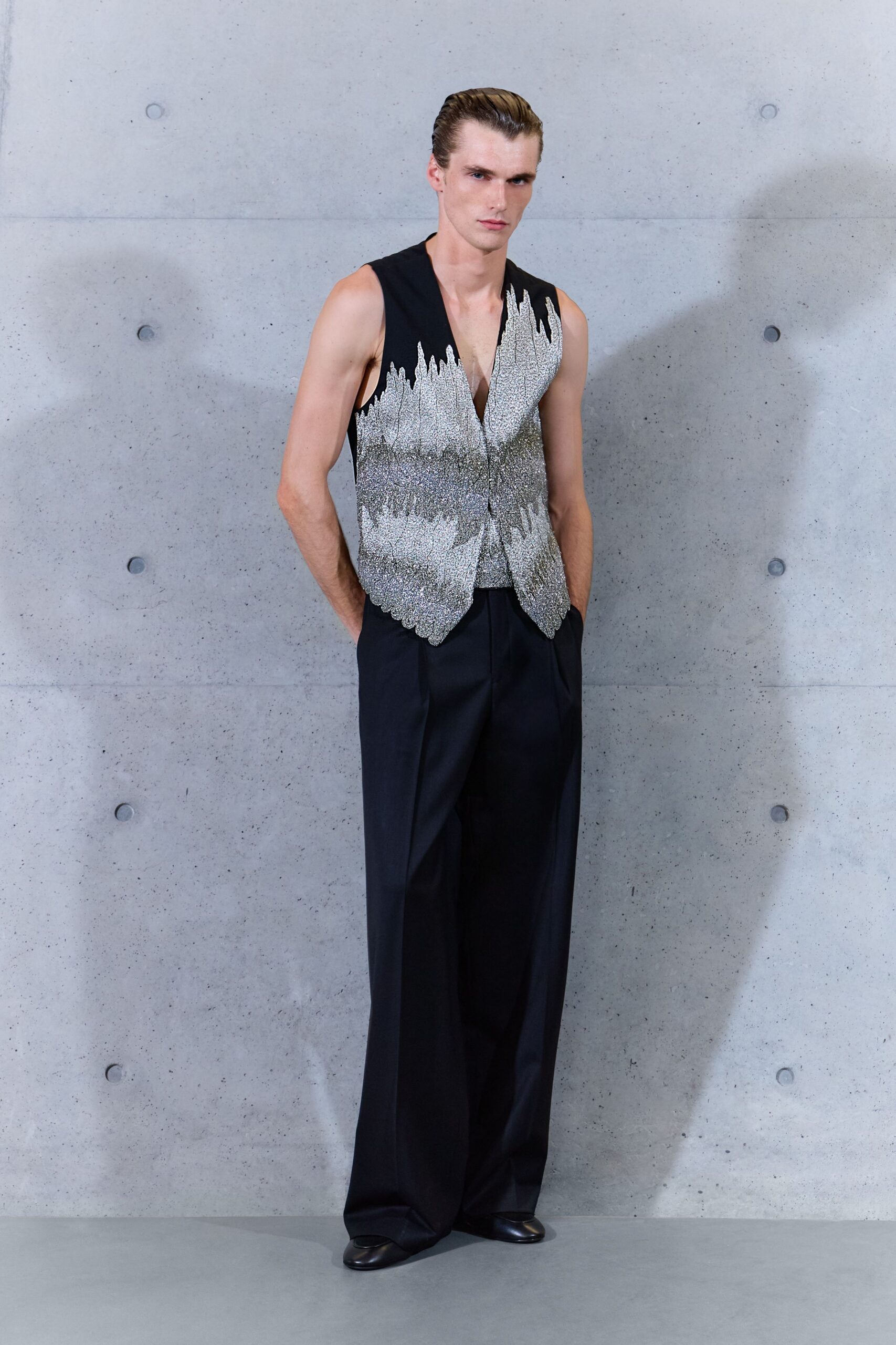 Sleeveless black waistcoat washed in silver beading, hem cut into scalloped points. Inky wide-leg trousers with pressed creases breaking softly over the shoe. Black leather square-toe shoes. Waiting by the museum's concrete stairwell after the benefit has thinned out.