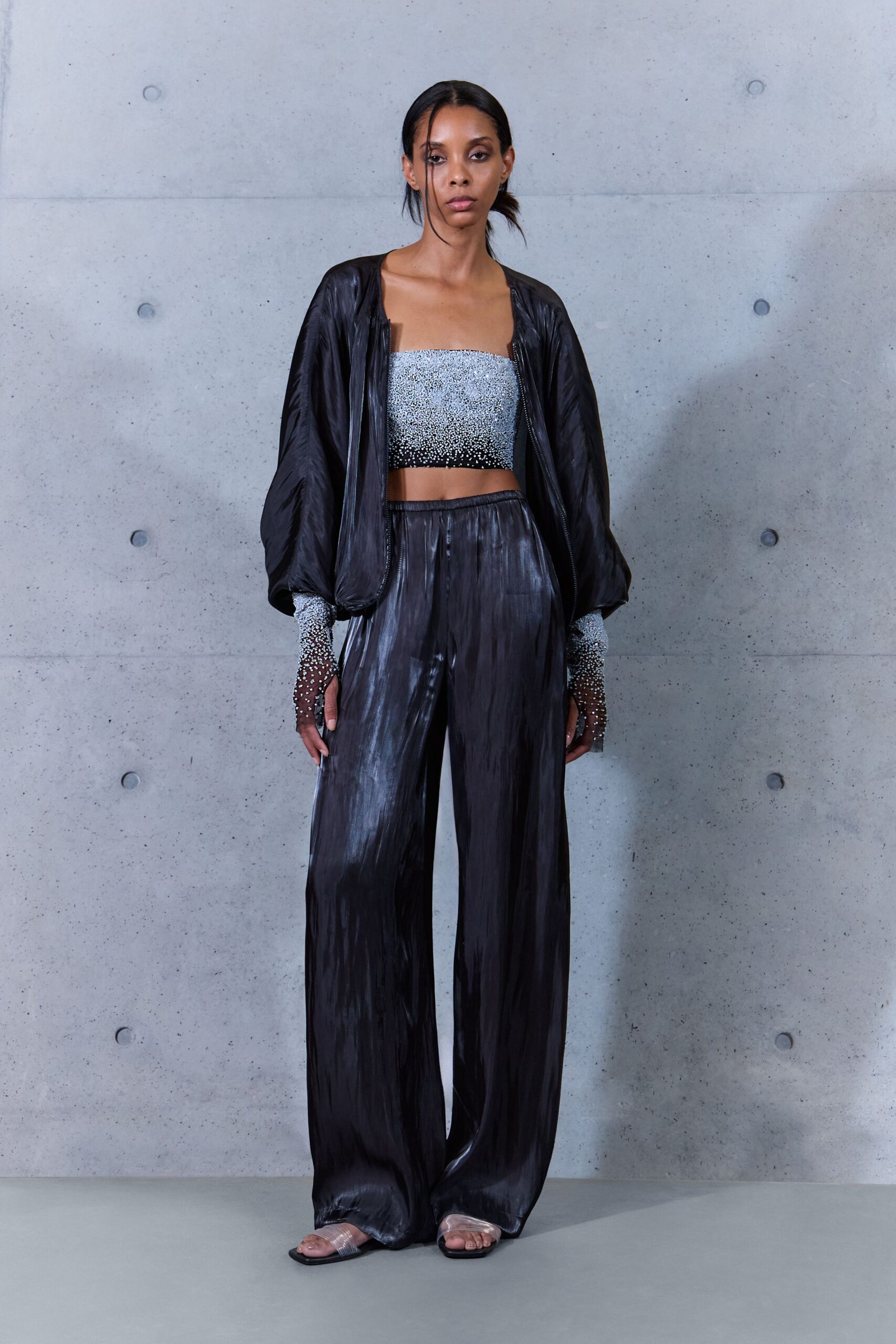 Black satin bomber, zip edges left hanging open and soft. Silver beaded bandeau fading to black at the lower edge. Matching crystal mesh gloves, stopping just below the elbow. Ink-shine wide trousers with a pressed vertical sheen. Clear strap slides, square black soles. Hotel corridor ice machine humming after midnight.