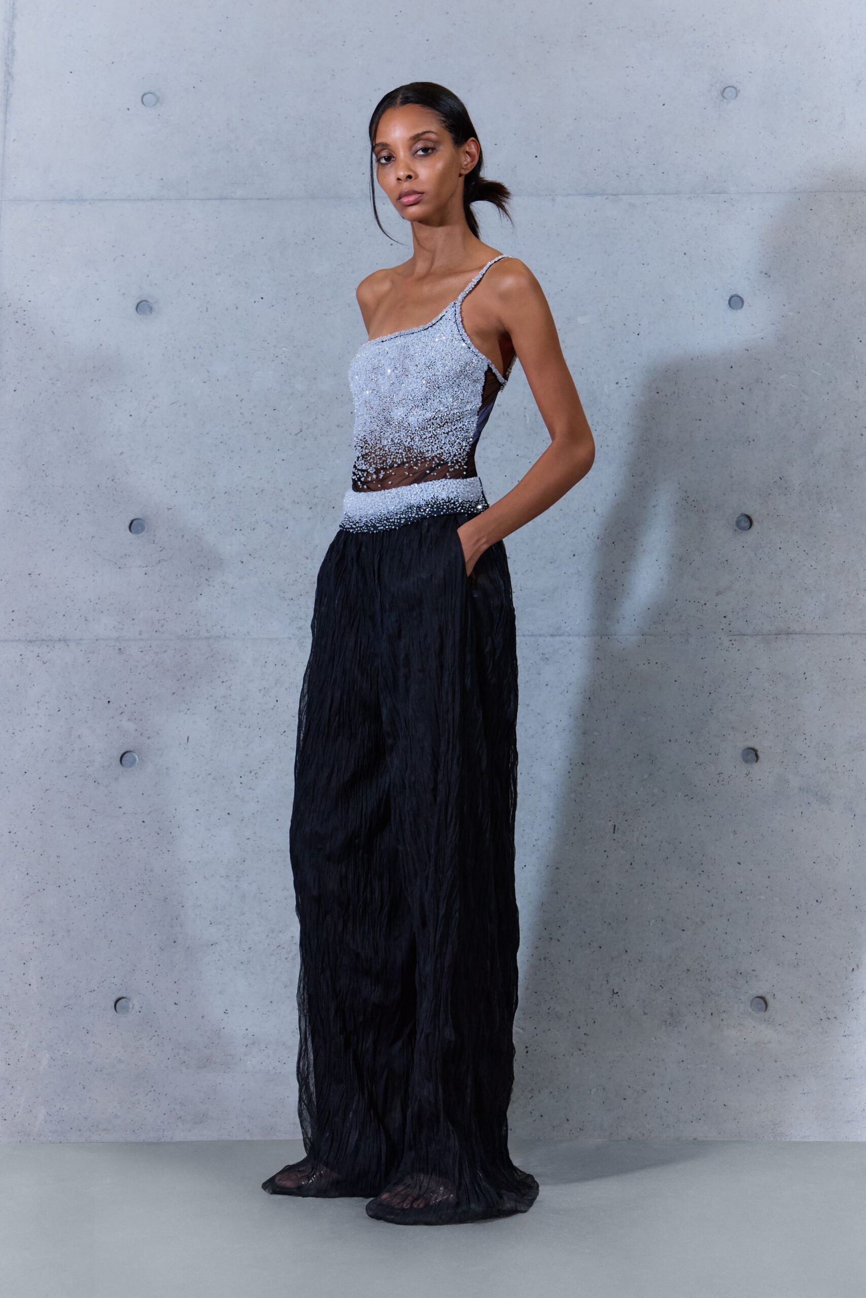One-shoulder evening dress, silver beading dissolving into sheer black mesh at the waist. Floor-length black crinkled chiffon skirt with a soft pooled hem. Black mesh shoes ghosting through the sheer hem. After midnight in a concrete lobby, waiting for the freight elevator.