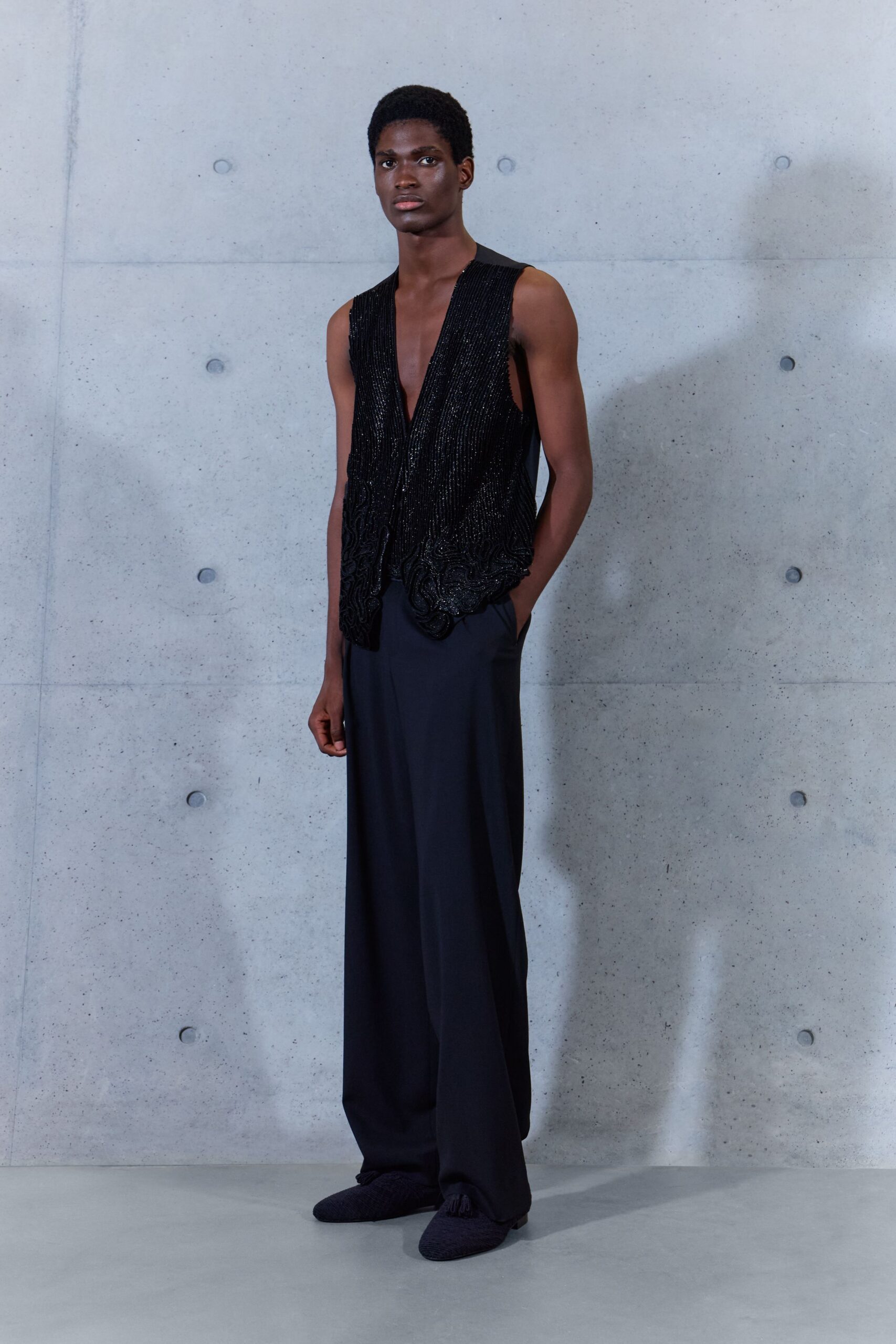Black beaded vest with a plunging V and scalloped embroidery at the hem. Ink-black wide-leg trousers in a fluid drape, pooling over the shoe. Dark woven slippers with a small tassel at the vamp. Hotel corridor after midnight, ice bucket sweating through a linen napkin.