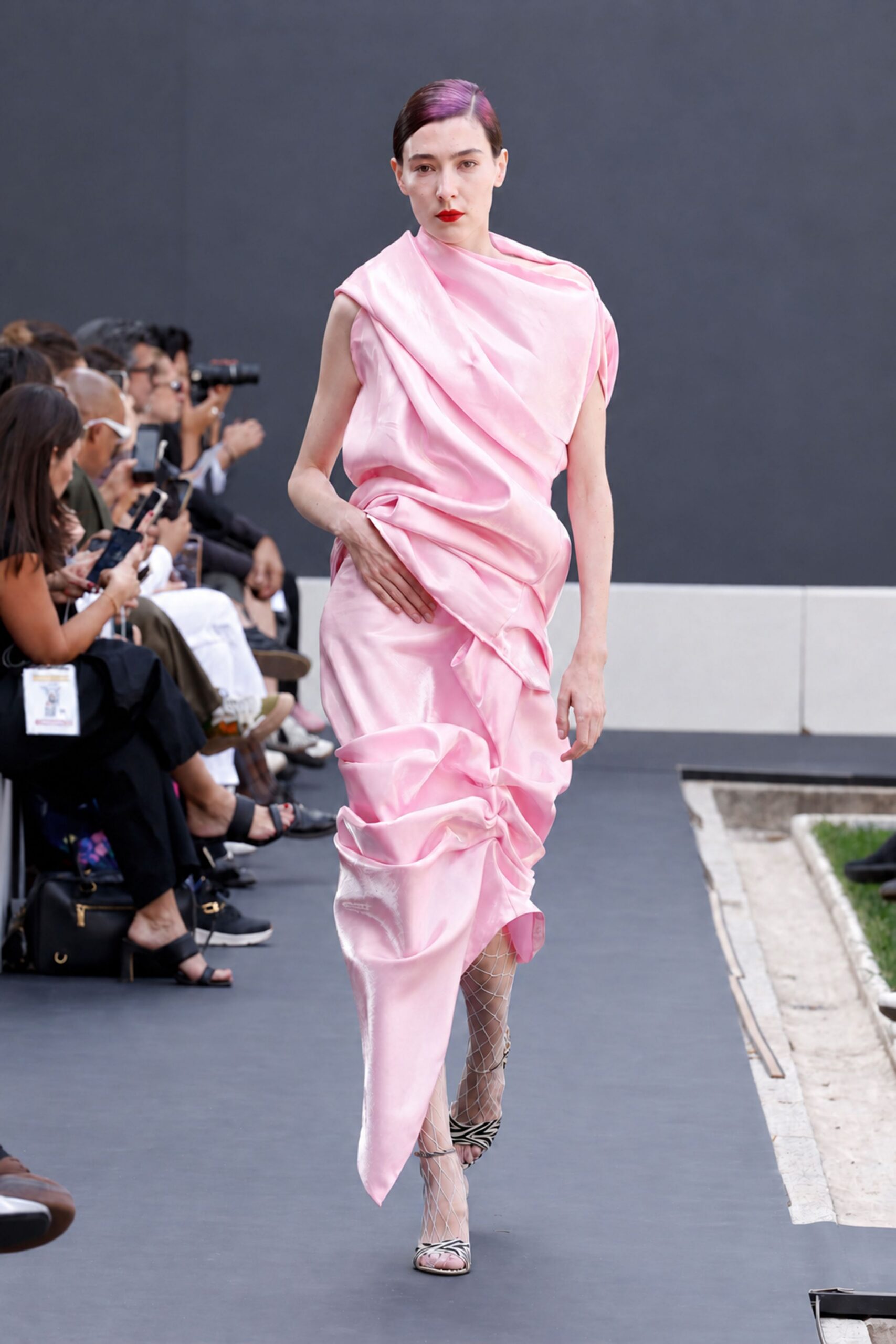 Blush satin dress draped from one shoulder into ruched, sculptural folds. White diamond-net stockings climbing past the calf. Zebra-print sandals with slim straps and a tiny ring buckle. Walking the museum courtyard after rain, flowers crushed in the wrapping paper.