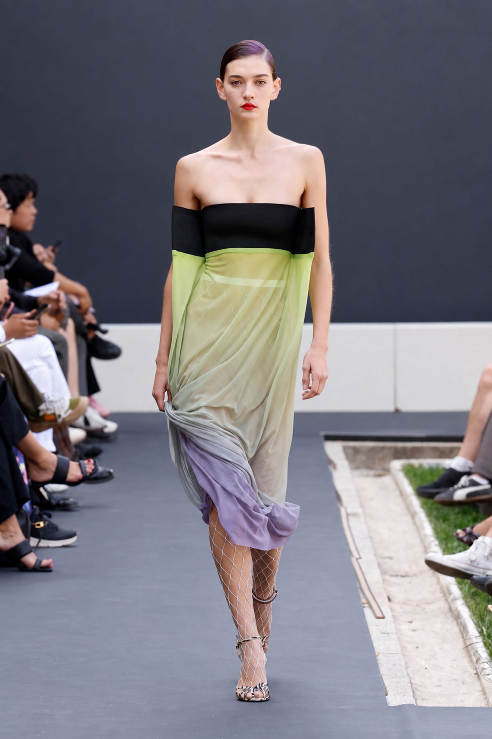 Black bandeau bodice with off-shoulder sleeves, chartreuse mesh draped to a lilac puddle hem. White diamond-net tights visible through the sheer layers. Ankle-strap peep-toes in a spotted hide pattern. Crossing a sculpture garden just after the lawn has been watered.