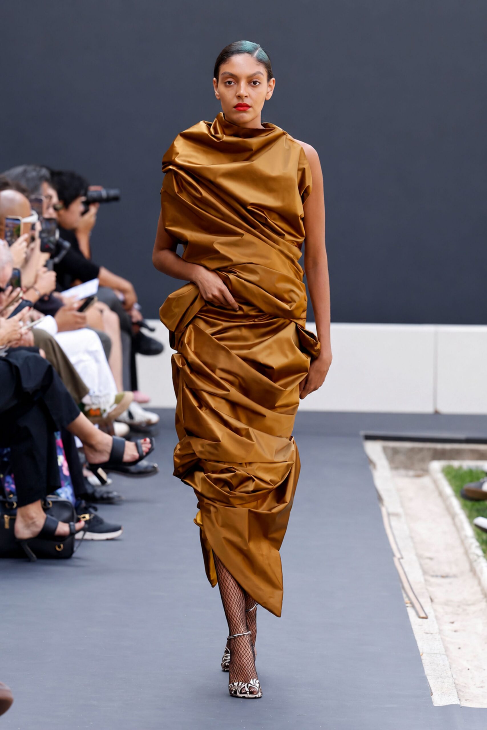 Bronze duchesse-satin dress, one shoulder bare and the whole length densely ruched. Black fishnet tights. Pointed pumps in swirling black-and-white print. Crossing a sculpture courtyard just before the auction starts.
