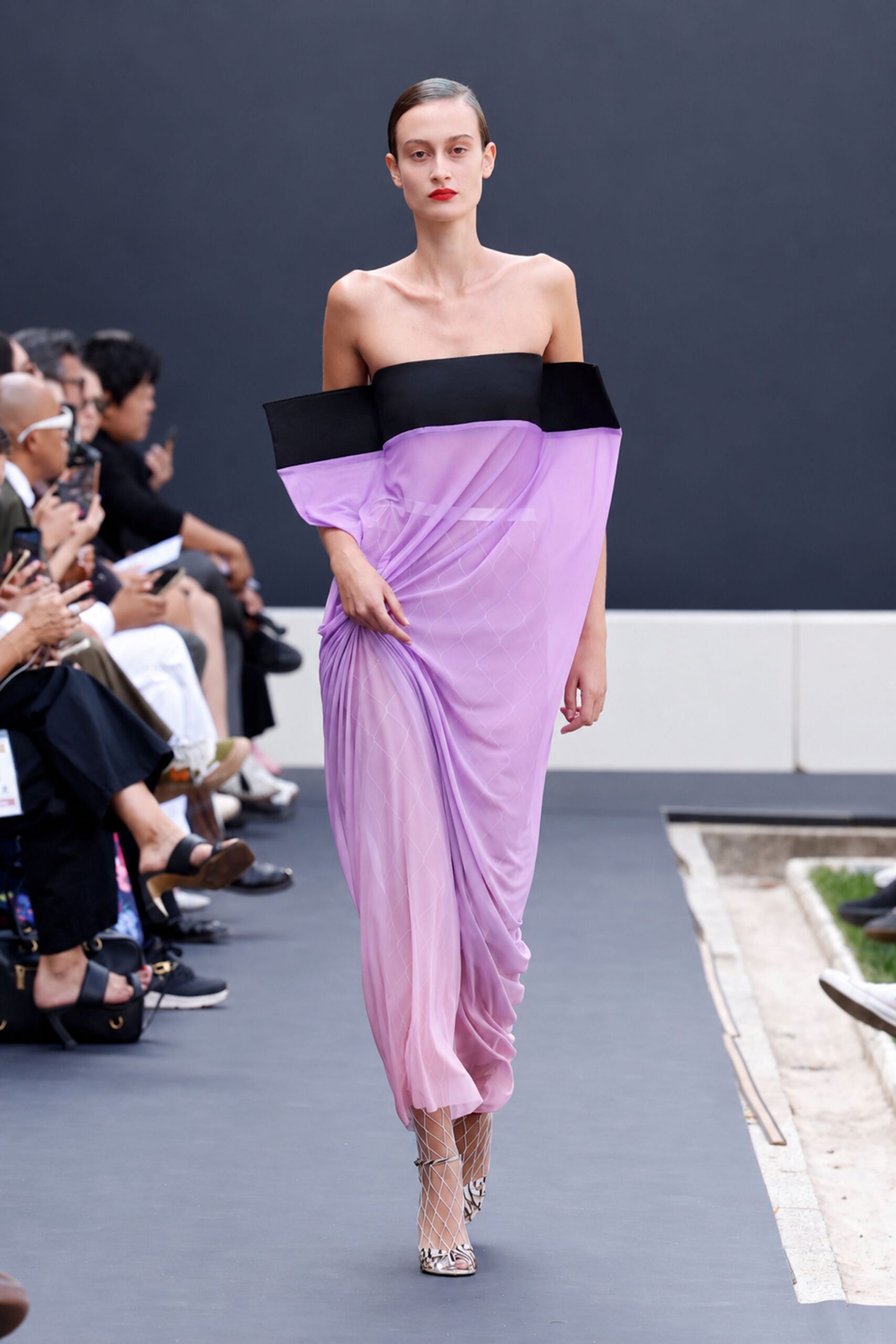 Lavender sheer draped dress with a black bandeau collar jutting off the shoulders. White diamond fishnet stockings visible through the gauzy skirt. Silver slingback heels with a black scribble print. Crossing a sculpture courtyard just before the museum lunch crowd spills out.
