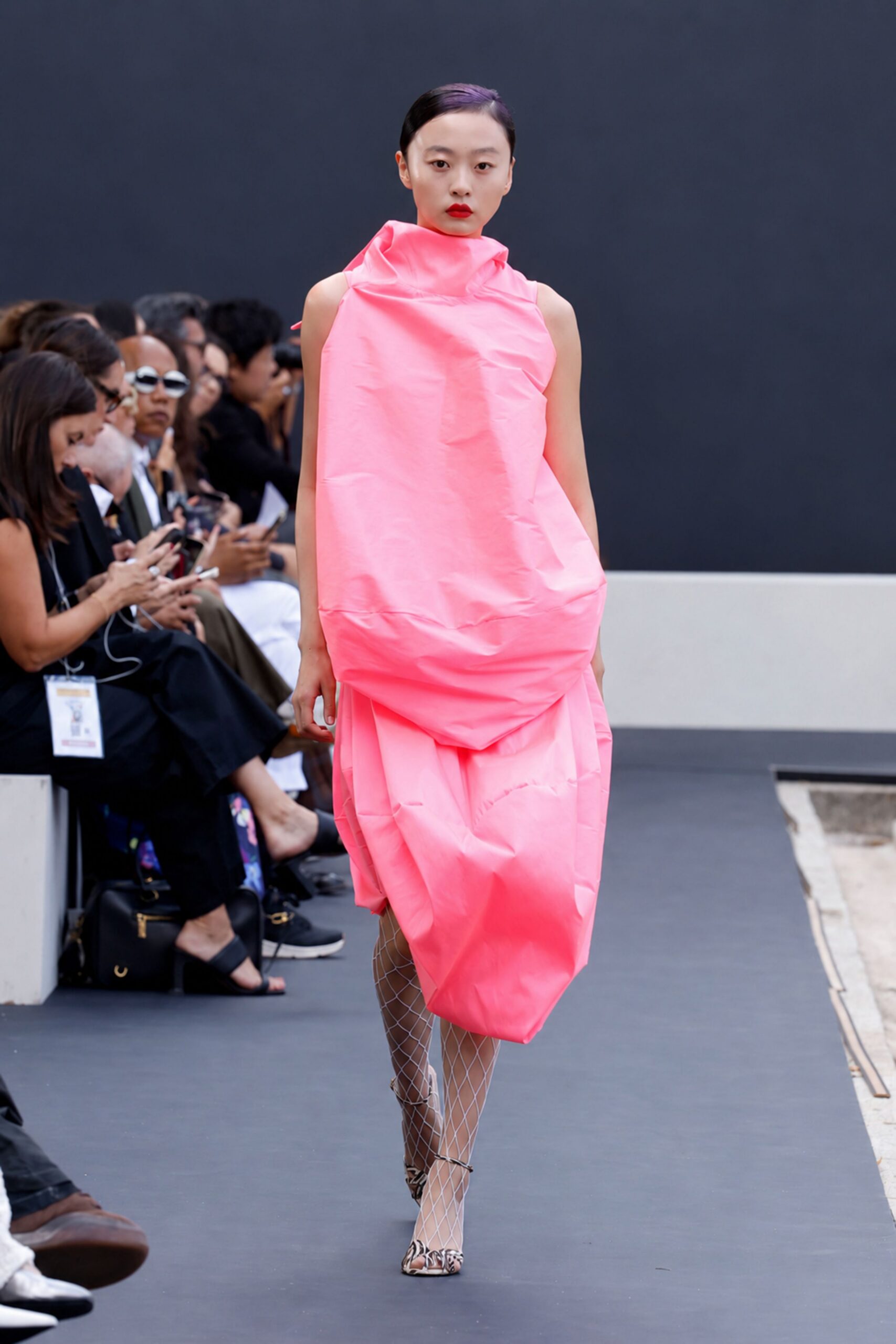 Neon pink taffeta cocoon dress, crumpled like tissue with a folded funnel neck. White diamond fishnet tights. Pointed pumps in a swirled brown-and-cream print. Crossing a sculpture court with wrapping still half on.
