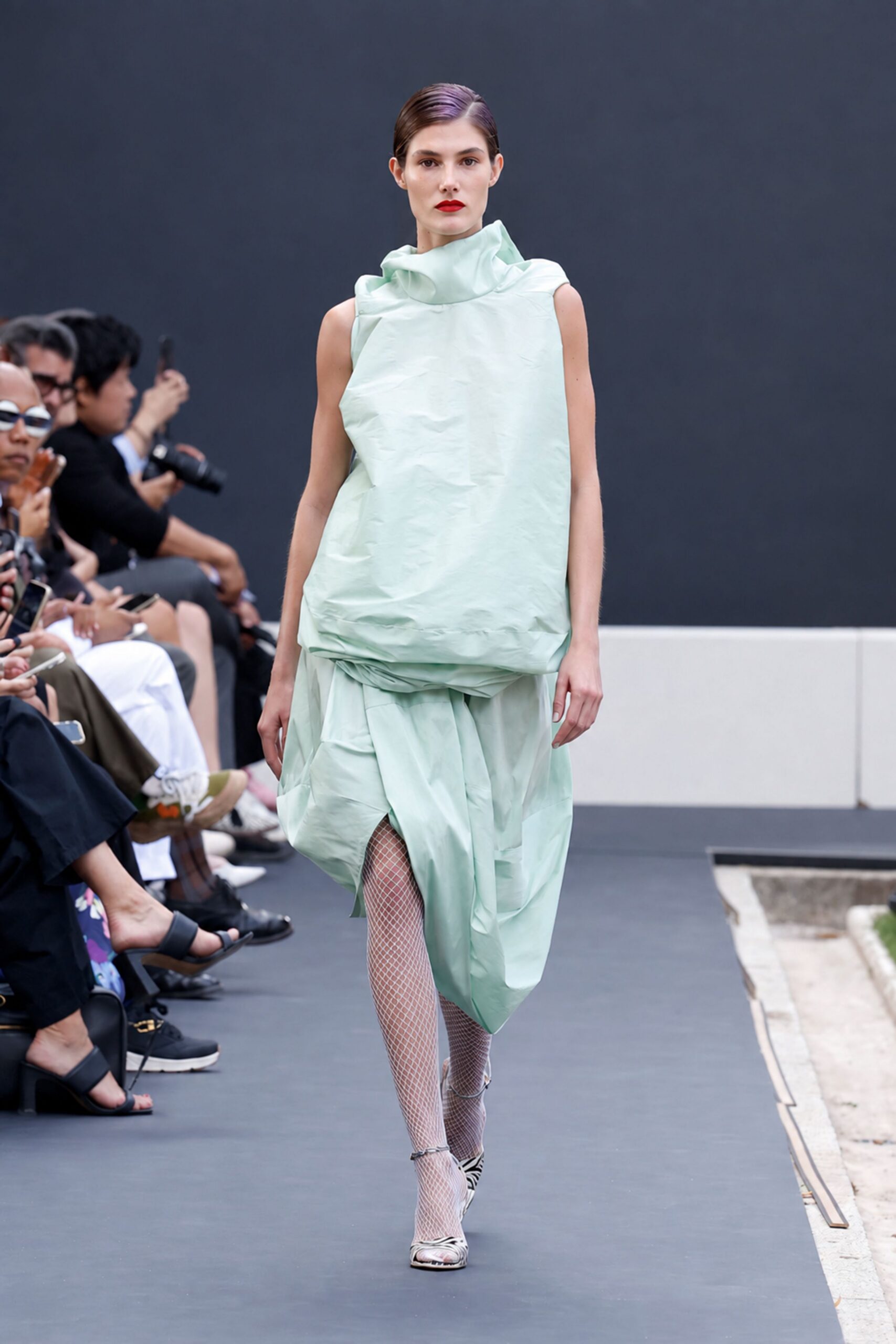 Mint paper-taffeta dress, cowl neck above a puffed rectangular torso. White fishnet stockings. Pointed zebra pumps with thin ankle straps. Crossing a sculpture garden before the fountains switch on.