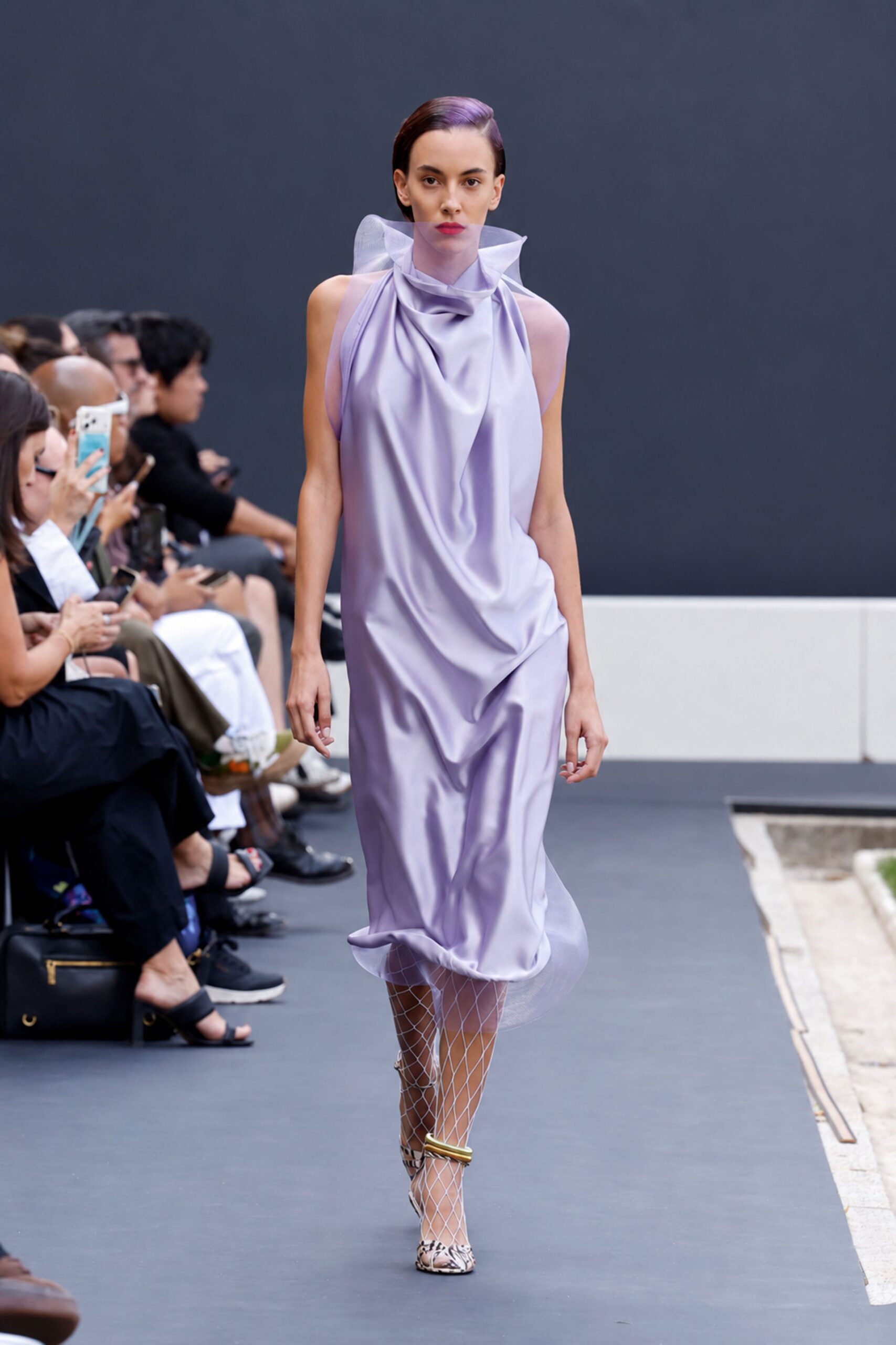 Lilac satin column dress, cowl neck rising into a sheer organza halo. White diamond-net stockings. Thick gold cuff anklet on one leg. Python pumps with a narrow pointed toe. Crossing a sculpture courtyard before the fountain switches on.