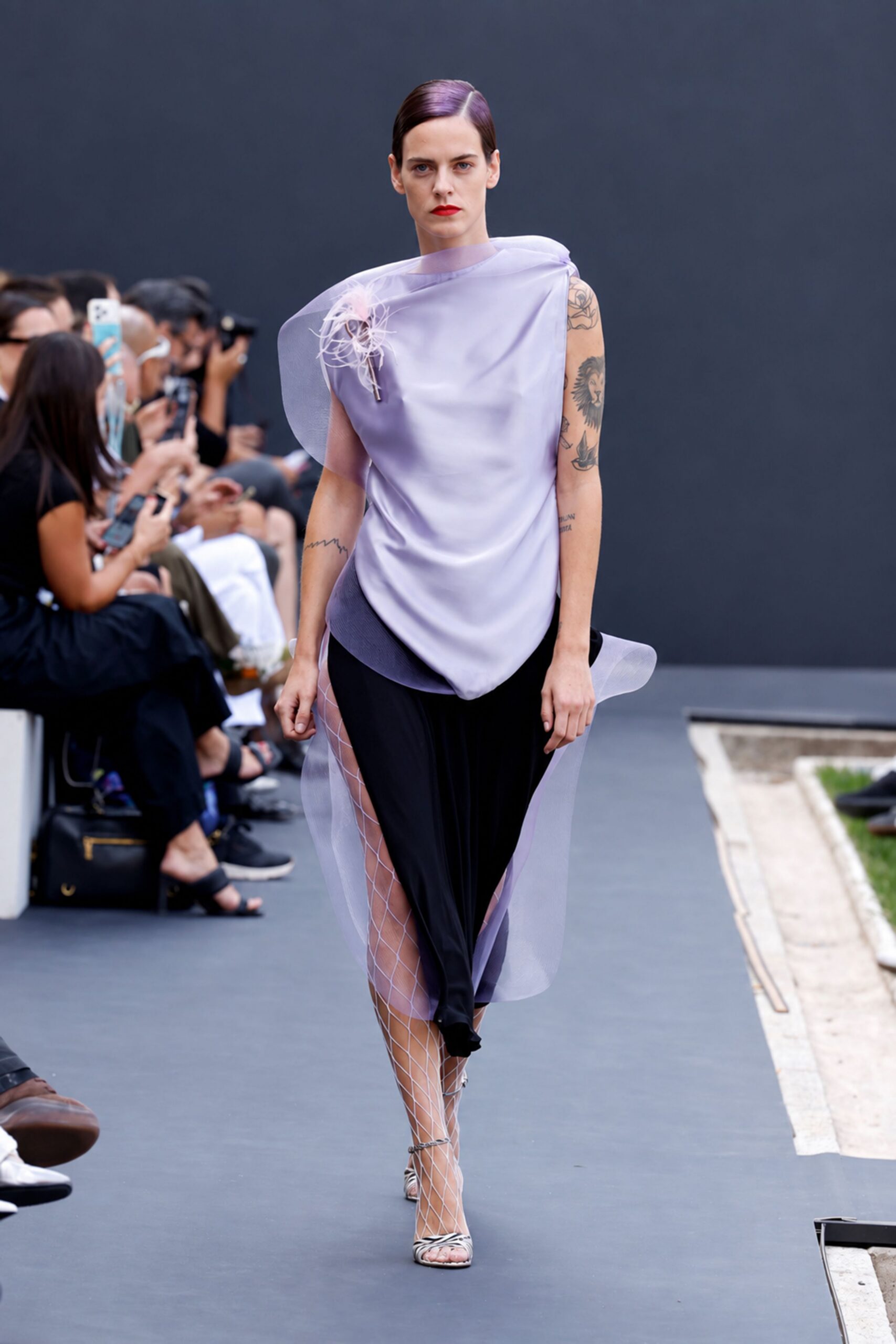 Lilac satin top draped askew under a sheer organza collar. Feathery pale bloom pinned high on the chest. Black jersey skirt split high, veiled with lilac organza panels. White diamond-net tights. Zebra-striped toe-ring sandals. Crossing a sculpture garden after rain, invitation folded in your fist.