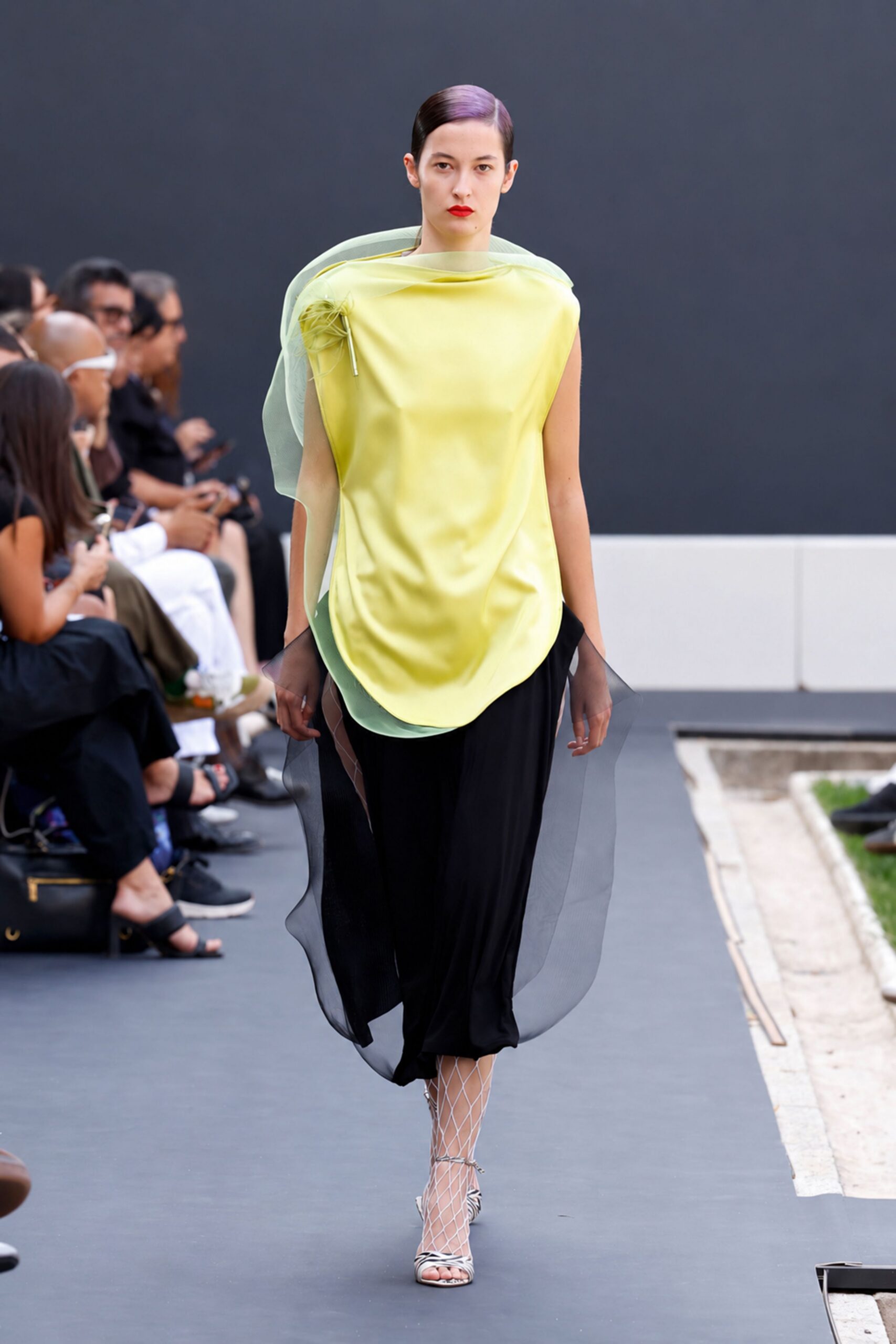 Butter-yellow satin tunic, draped inside a pale mint organza shell. Spiky rosette brooch pinned high on the left shoulder. Black fluid skirt, overlaid with sheer smoky panels that ripple. White diamond-net stockings. Zebra-print ankle-strap sandals. Crossing a sculpture garden before the heat leaves the paving stones.