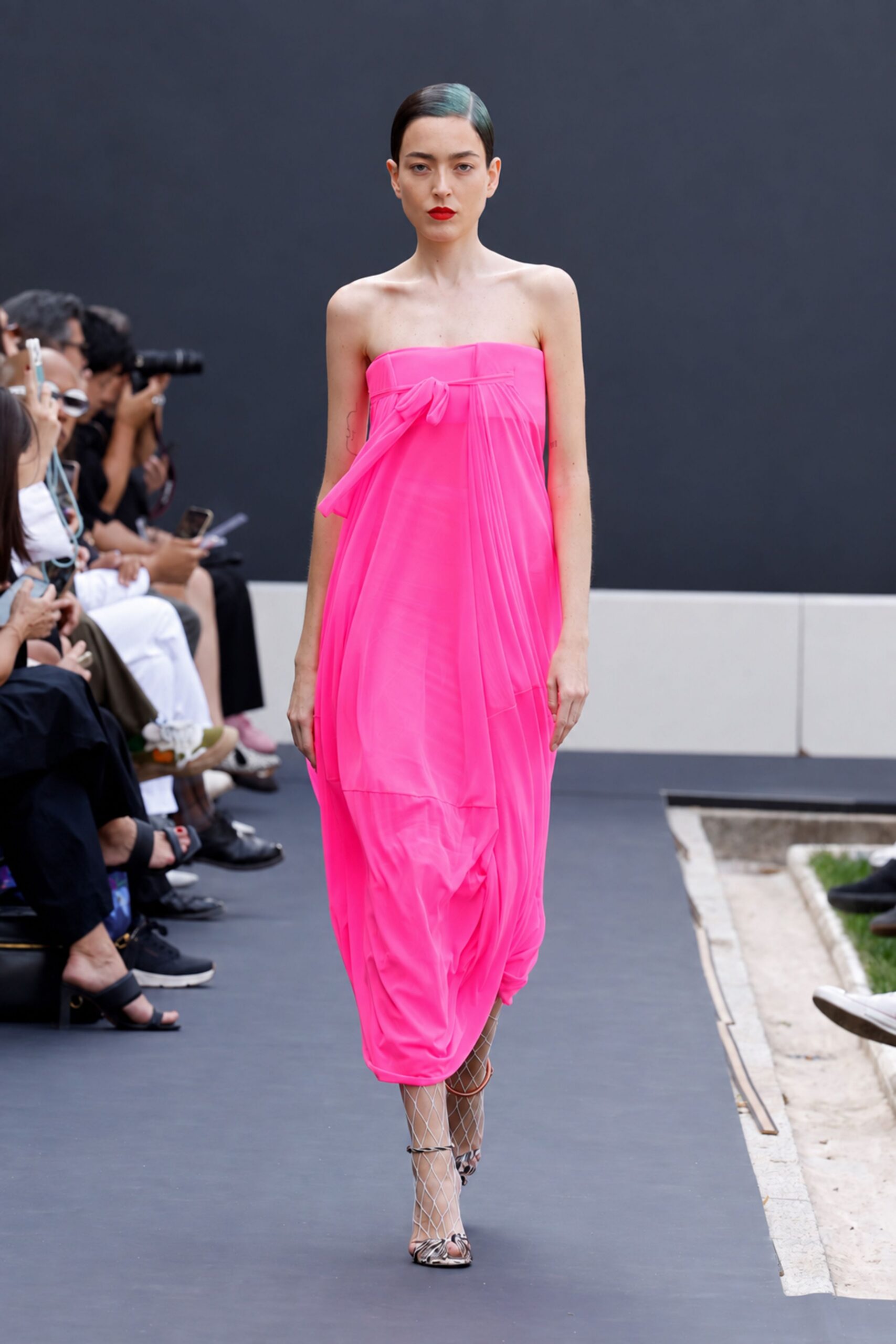Neon-pink strapless column, sheer panels pleated into a drooping side bow. Python-print sandals wrapped with white diamond-net ankle sleeves. Crossing a sculpture courtyard at noon with lipstick still too fresh.