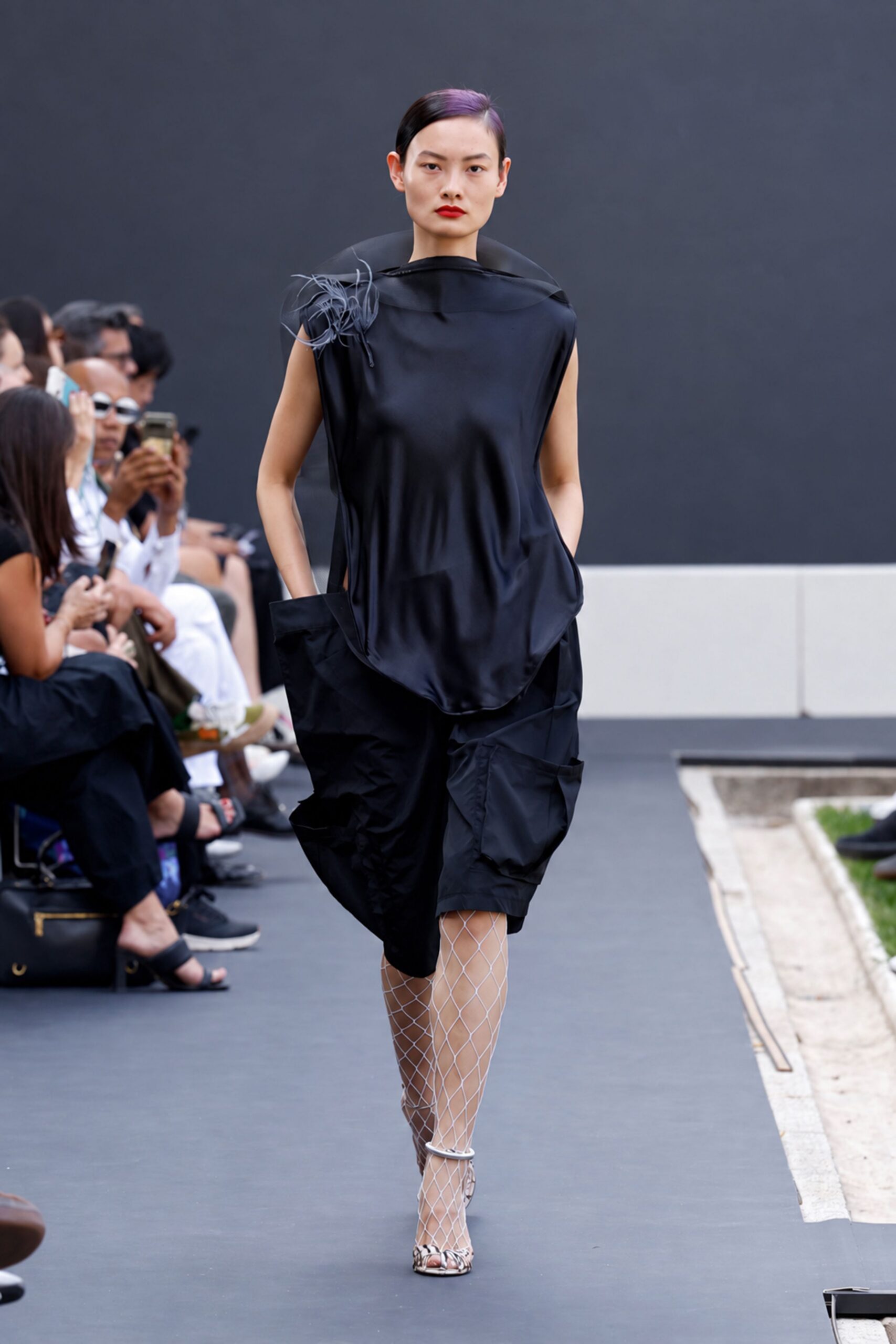 Black satin tunic with a sheer curved collar and wispy shoulder plume. Black balloon shorts in crisp technical cloth, side pockets jutting outward. White diamond fishnet tights. White ankle-strap heels with snakeskin toes. Crossing a museum courtyard just before the storm finally breaks.