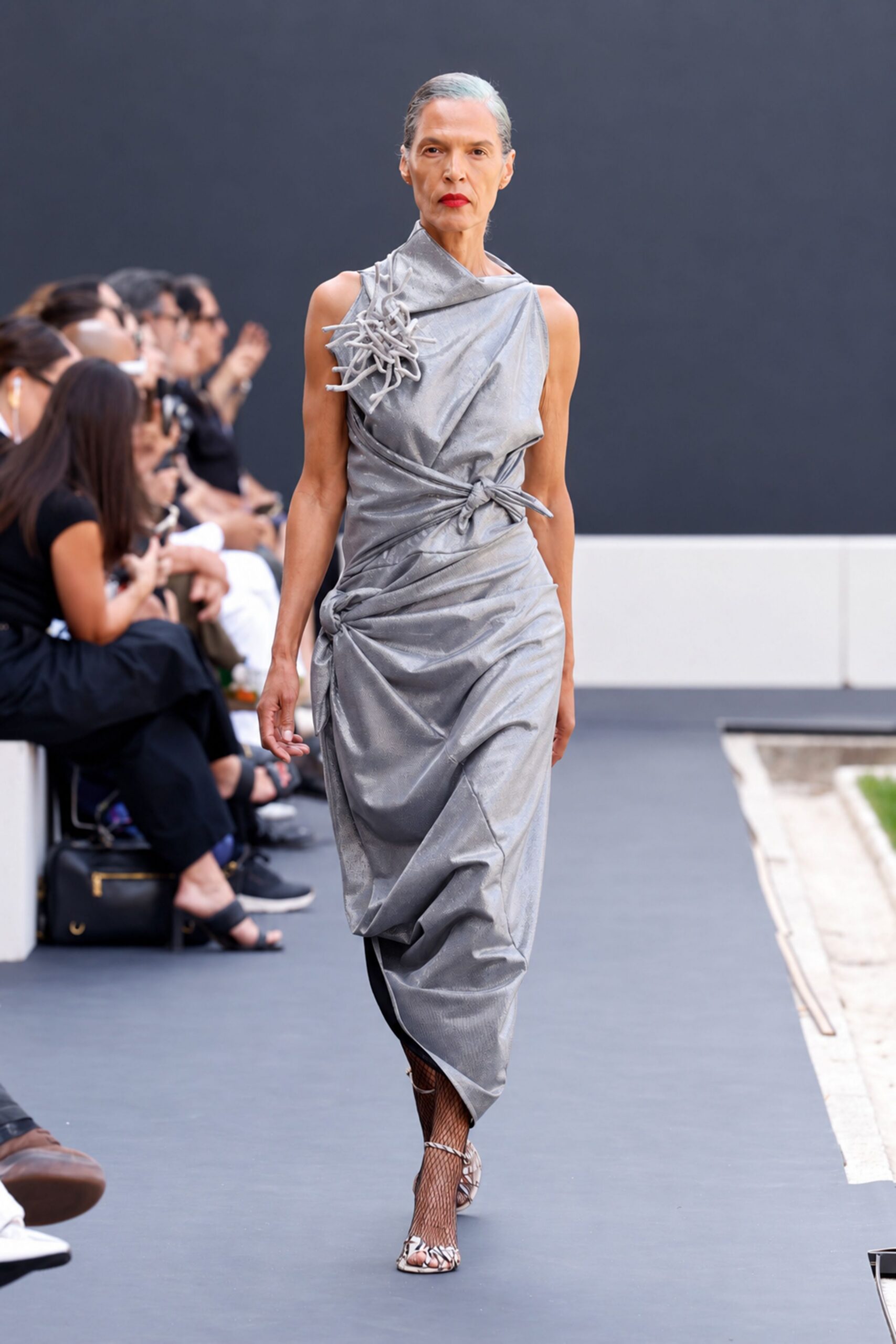 Silver draped dress in crinkled foil jersey, knotted at hip and waist. Tangled tubular rosette climbing one shoulder. Black fishnet to the knee. White caged sandals laced over the toes. Crossing a museum courtyard just after the fountain has been turned off.