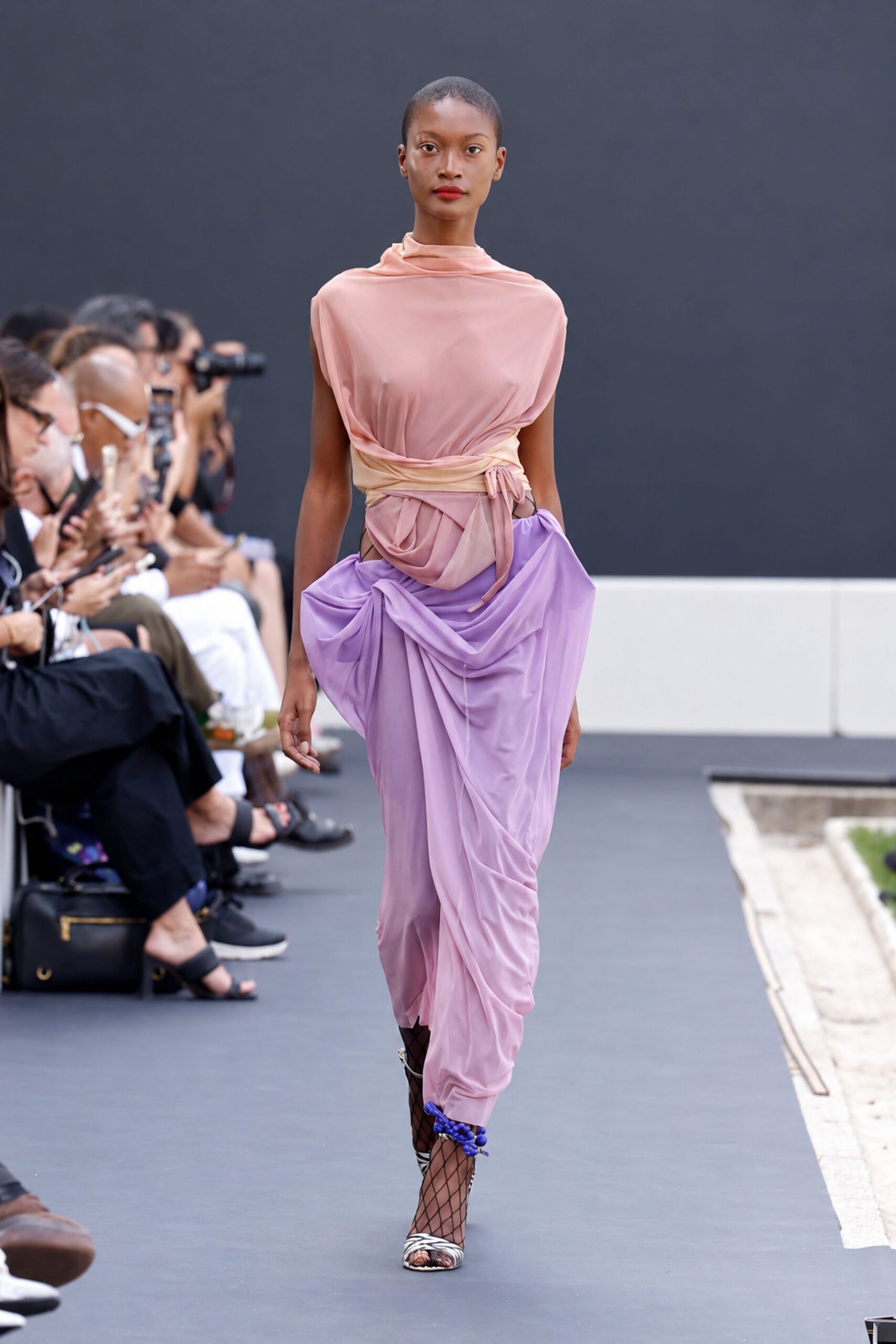 Blush sheer jersey draped into a high cowl and twisted waist wrap. Lilac jersey harem trousers, pouched at the hips and ruched to the ankles. Black diamond-net tights. Zebra-print sandals with a clustered violet ankle tie. Crossing a hotel terrace after the pool has emptied but the heat has not.