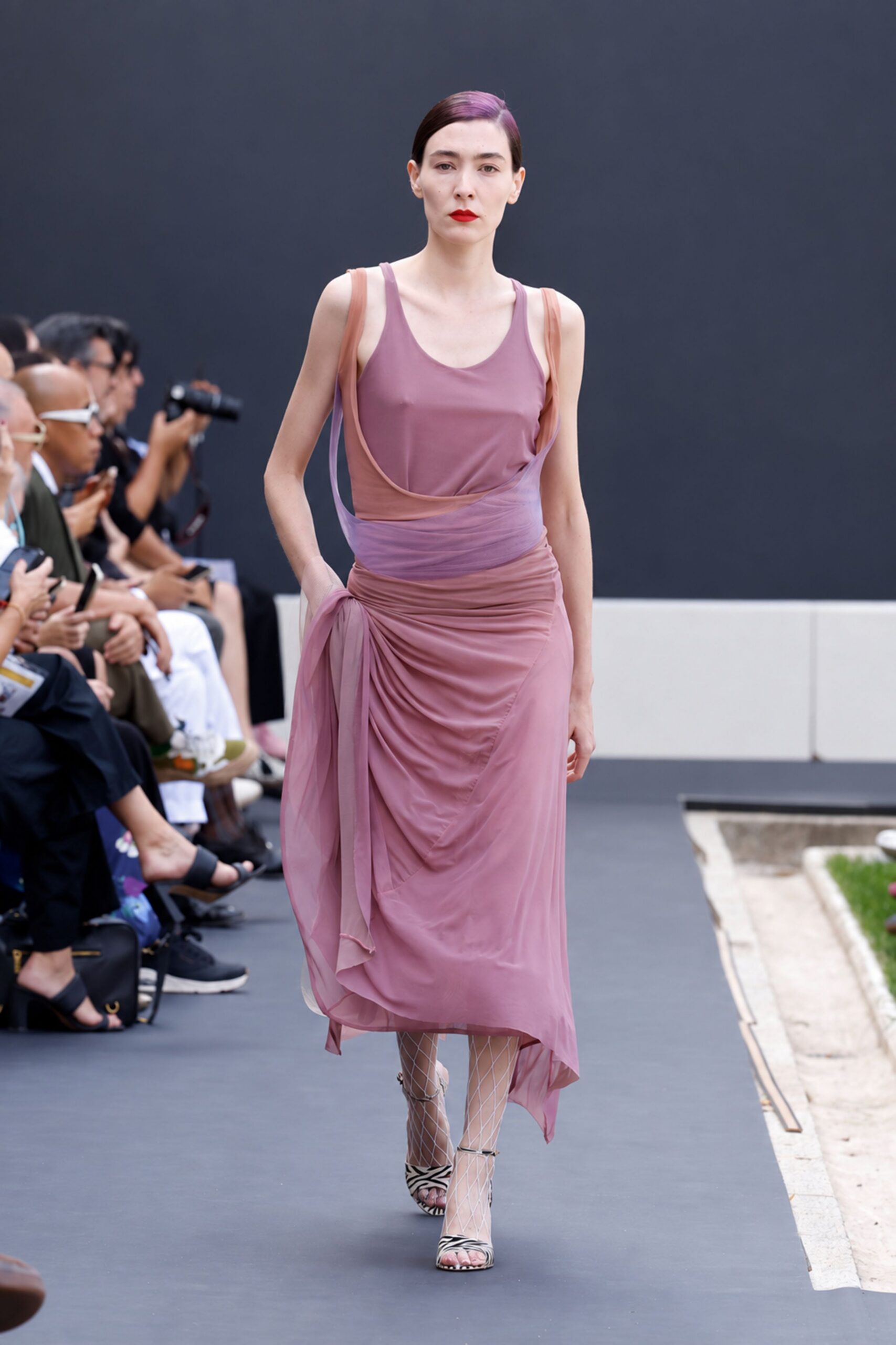 Dusty mauve jersey slip dress with a scooped neckline. Blush chiffon wrap overlay, ruched low on one hip with trailing ties. White diamond fishnet tights. Zebra-print ankle-strap sandals. Crossing a sculpture garden after the wedding, grass clippings on the path.