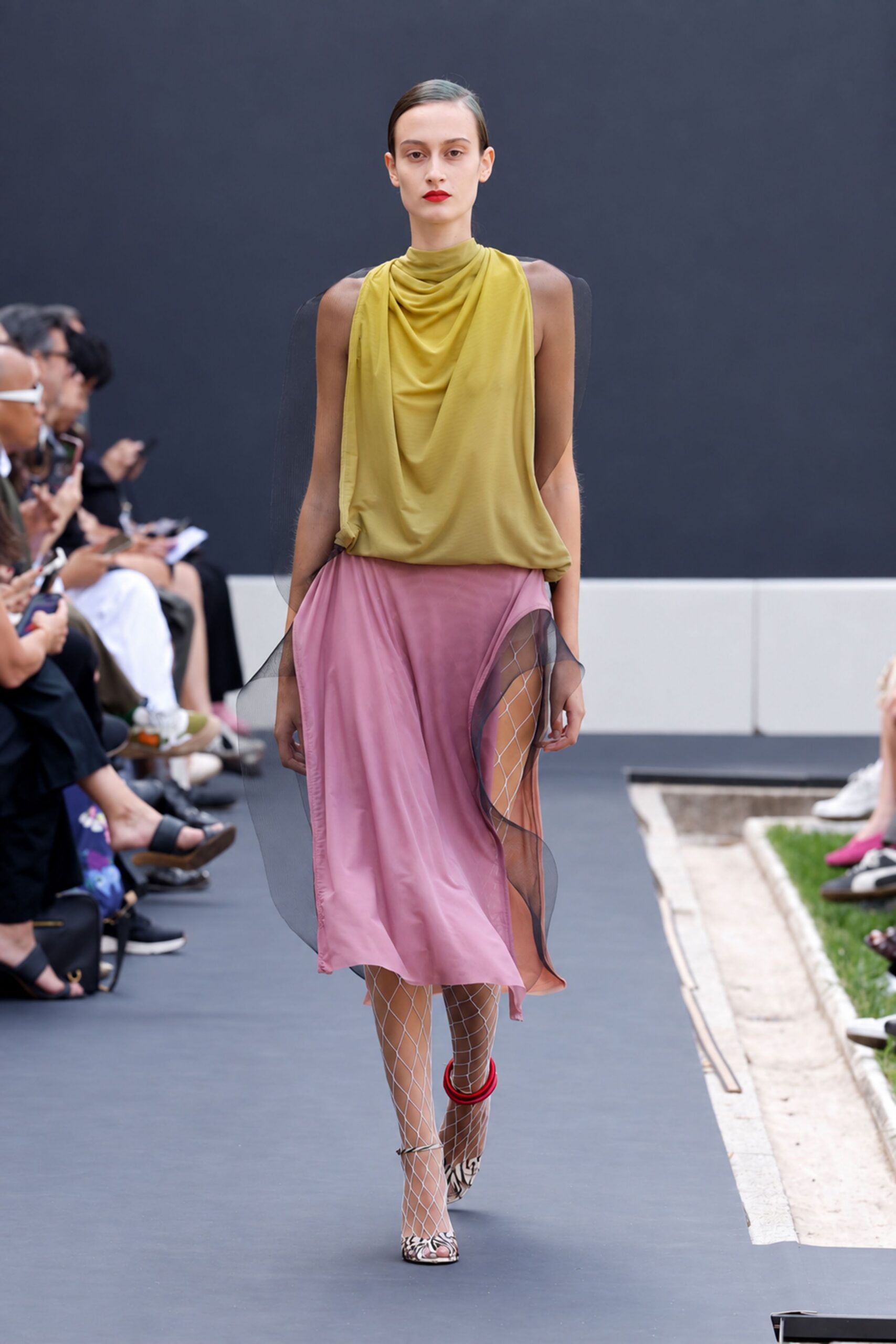 Olive jersey top draped into a cowl from a close mock neck. Smoke organza panels float loose at the sides. Dusty rose slip skirt cut with a high curved front slit. White diamond-net tights. Zebra peep-toe pumps with a red ankle ring. Crossing a sculpture garden after rain, dress code only half obeyed.