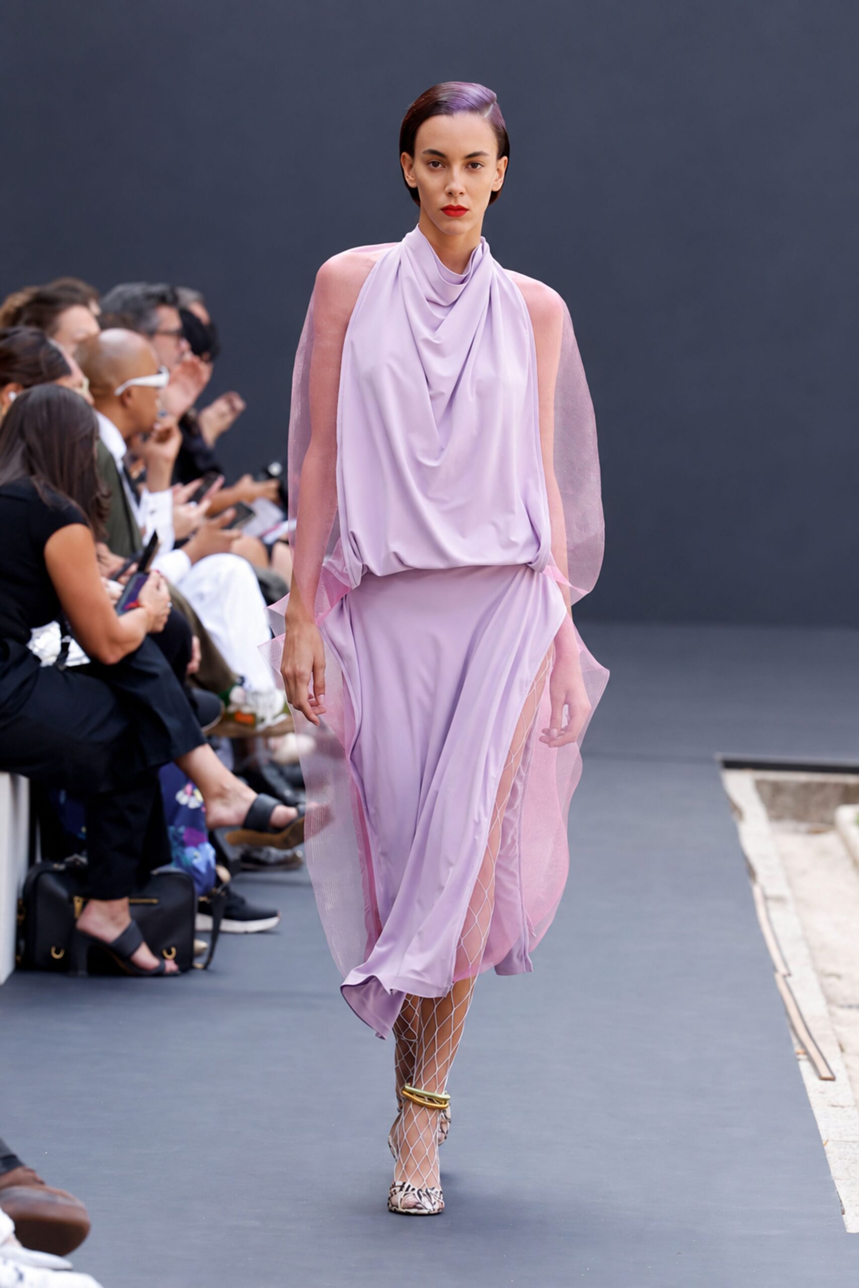 Lilac draped jersey dress with a cowl neck and sheer pink organza panels. White diamond fishnet stockings climbing above the slit. Python peep-toe heels with stacked gold rings at the ankle. Crossing a museum courtyard just after rain, carrying nothing but lipstick.