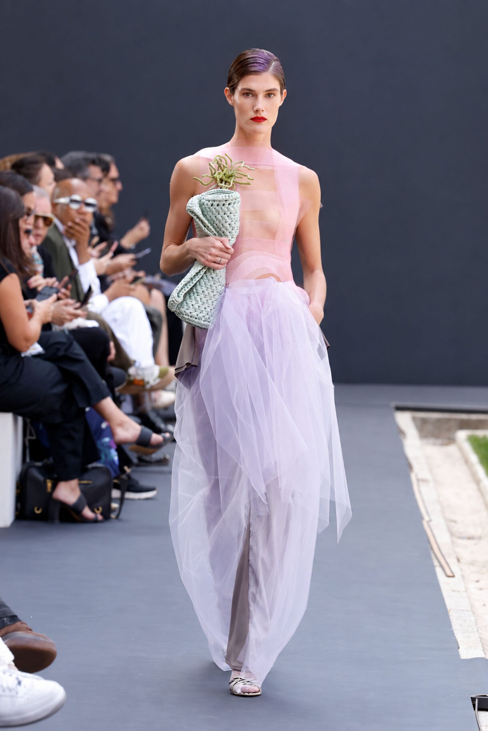 Blush sheer bands wrap the torso, one strip skewed off the shoulder. Taupe liquid-satin trousers veiled by lilac tulle ballooning from the hips. Mint chunky-woven clutch topped with wiry green tendrils. White cage sandals over pale fishnet. Crossing a sculpture garden after rain, still carrying the florist paper.