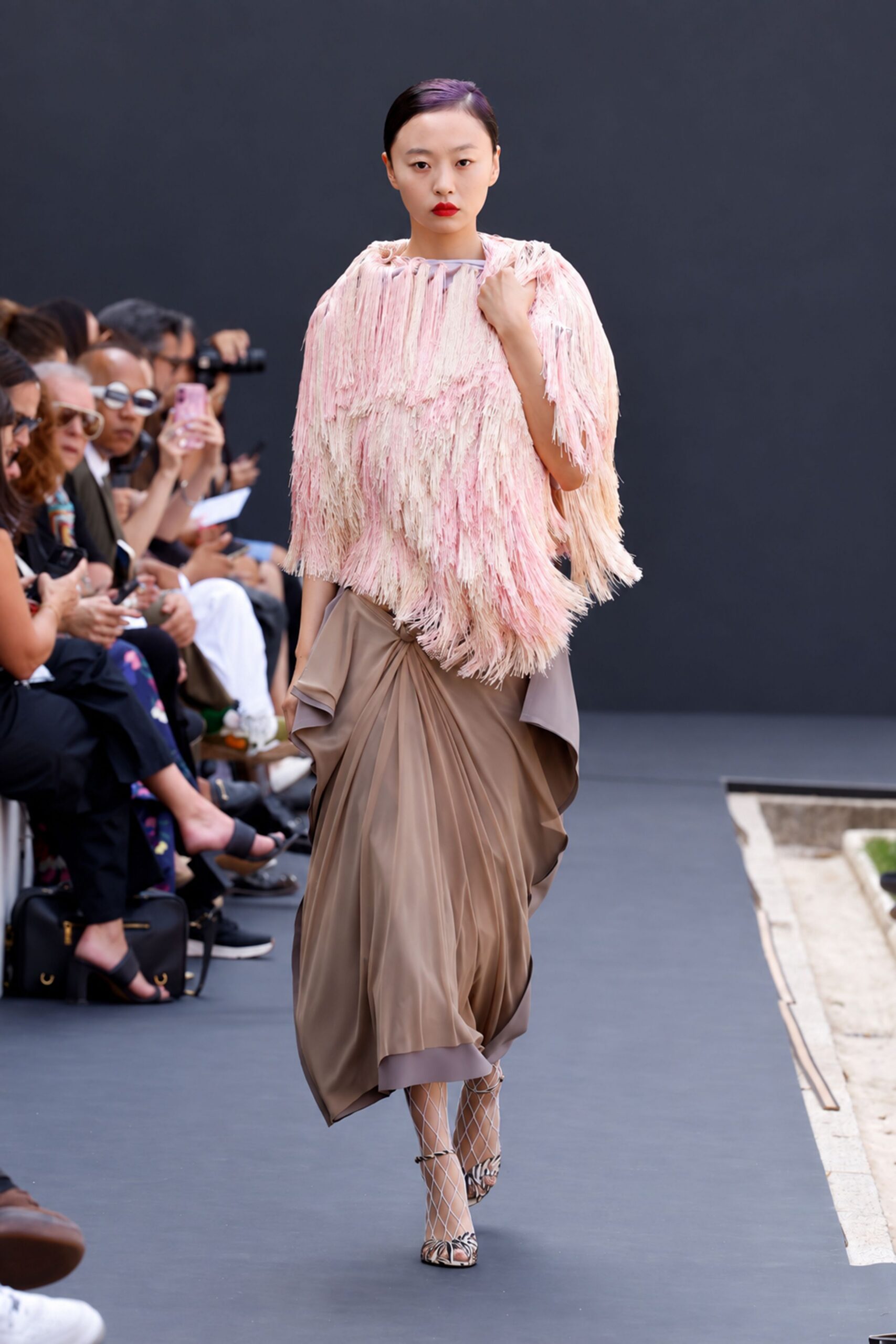 Blush and cream fringe layered in dense bands from neck to hem. Taupe draped skirt with a gathered knot and lilac underlayer peeking below. White diamond fishnet tights. Zebra-print peep-toe heels with lattice straps over the foot. Crossing a museum courtyard after rain, bouquet paper still in your hand.