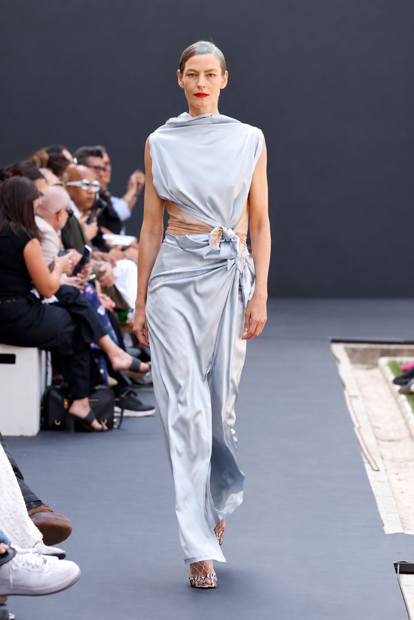 Pearl-grey satin draped from a cowl neck into side-tied folds. Nude stretch mesh cinching the waist beneath the knot. Snakeskin peep-toe heels with white lattice over the foot. Crossing a museum courtyard just before the fountain switches on.