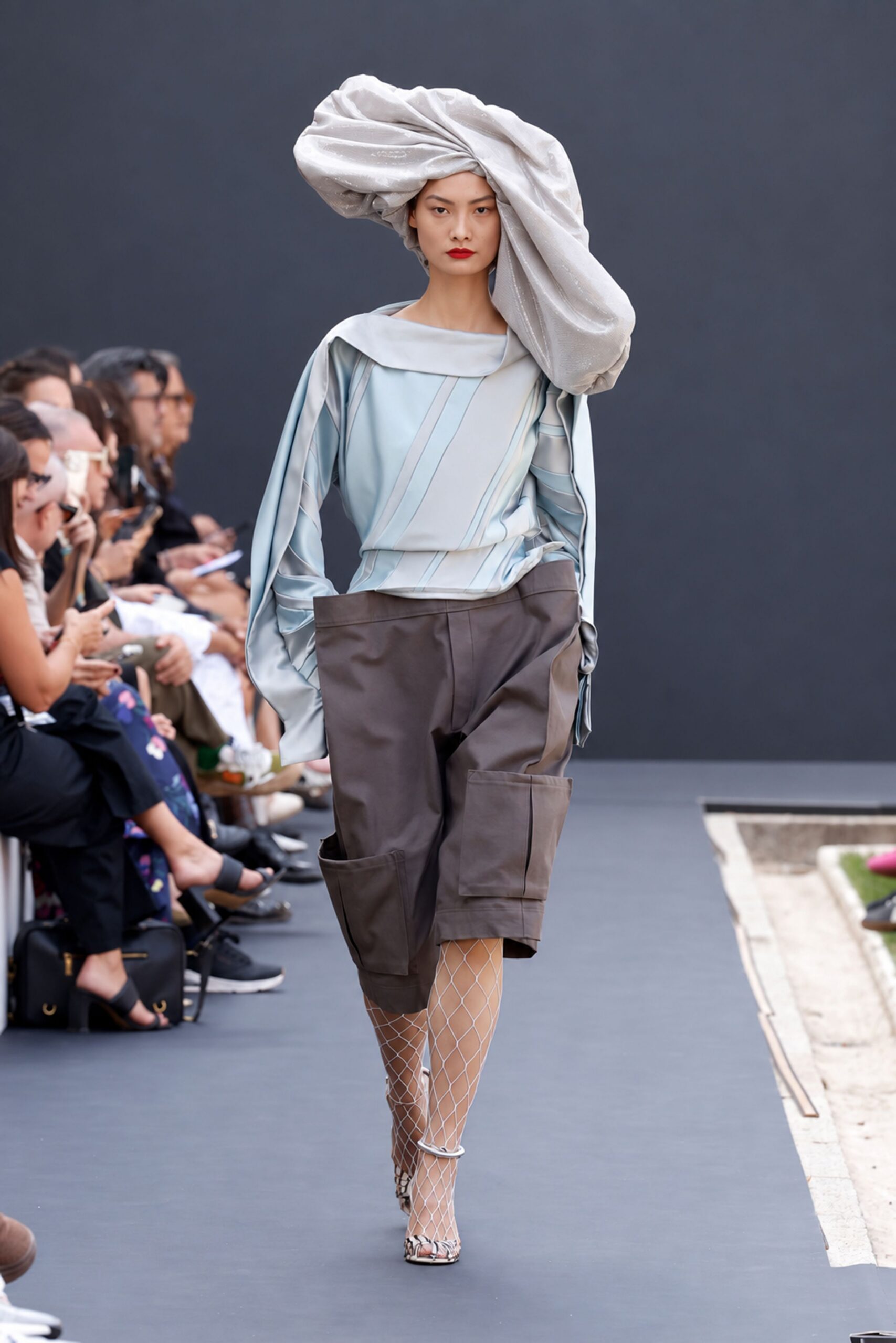 Huge silver lamé headwrap twisted into a lopsided cloud. Pale ice satin blouse with slashed sleeves and a folded boatneck. Taupe knee-length cargo shorts with oversized patch pockets jutting outward. White diamond fishnets. Snake-print ankle-strap heels. Crossing a sculpture courtyard before the fountain switches on.