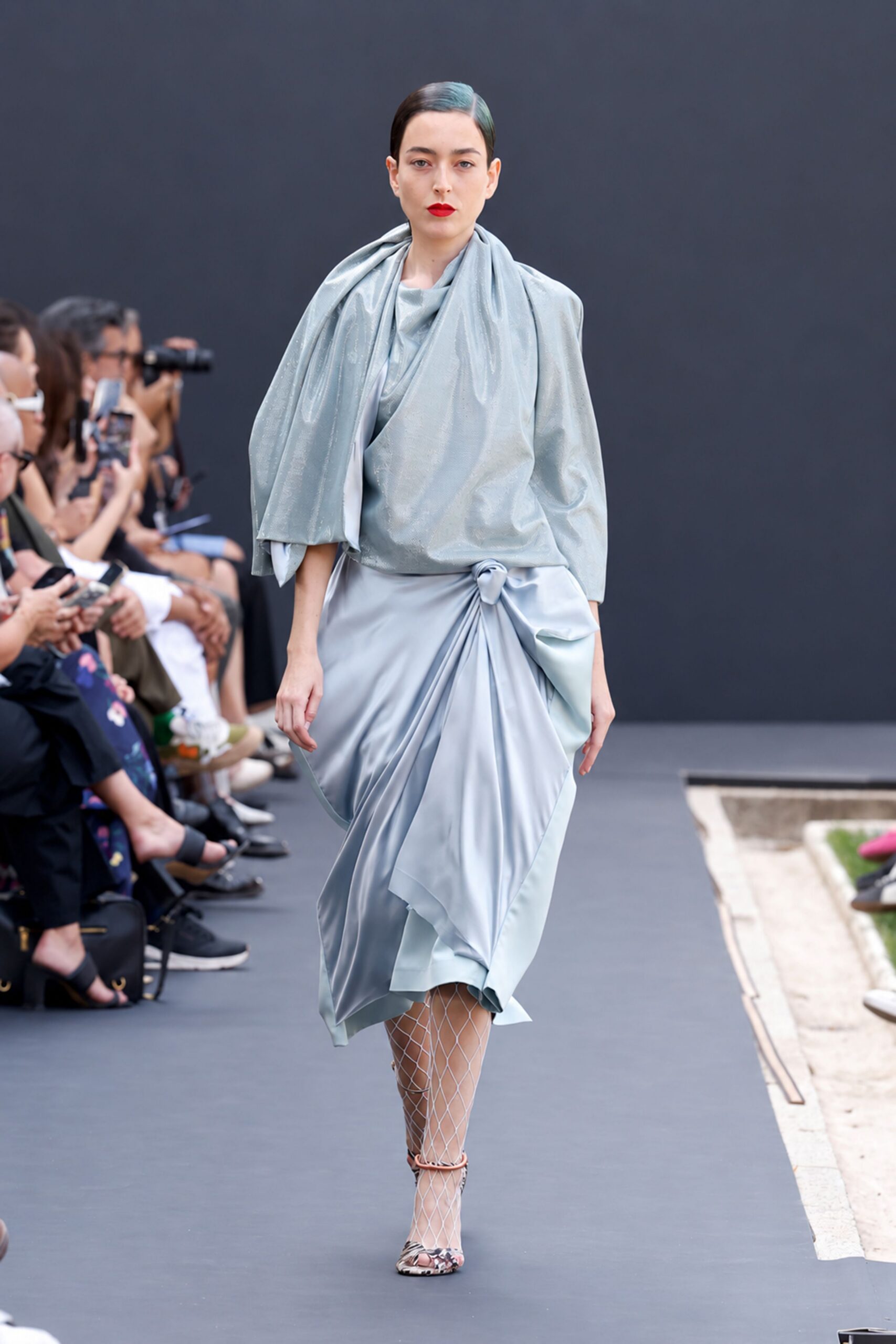 Pale ice-blue lamé top with a scarf panel slung off one shoulder. Matching satin skirt ruched into a knot at the right hip. White diamond-net tights. Snakeskin peep-toe heels with a blush ankle strap. Crossing a museum courtyard after rain, invitation folded in one hand.