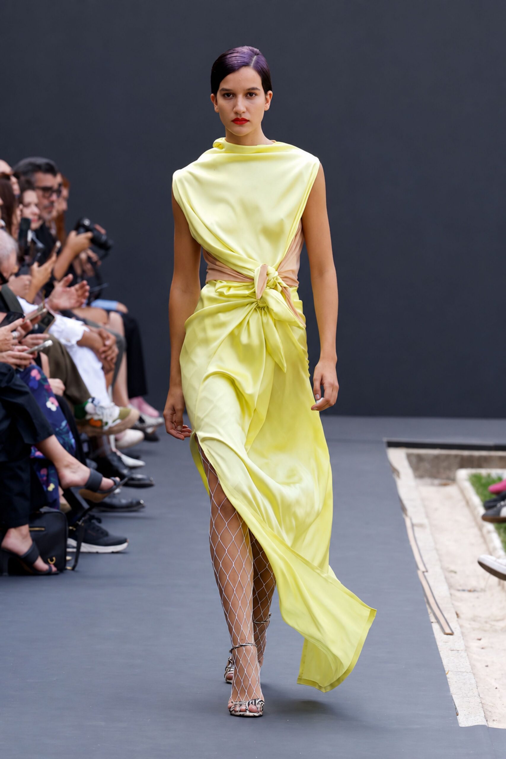 Lemon satin dress draped into a bloused cowl and knotted waist. Nude mesh side panels cinching the torso under the wrap. White diamond-net stockings climbing high through the front slit. Snake-print heeled sandals with thin straps over the toes. Crossing a sculpture court just before the fountains switch on.