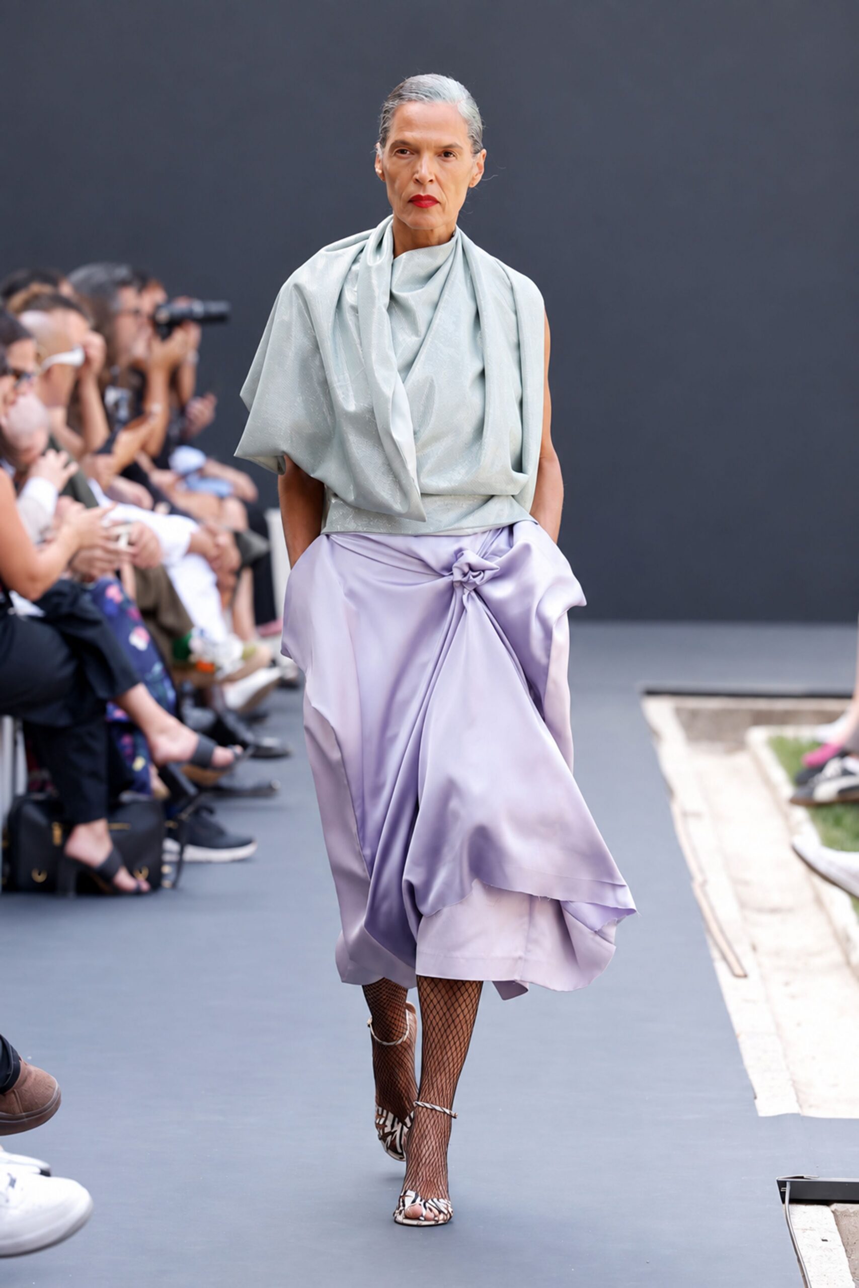 Pale celadon lamé top, draped into a soft cowl and scarf fall. Lilac satin wrap skirt knotted high at the hip, hem pooling unevenly. Black fishnet tights. Cream lace-up heels in a cutout zebra pattern. Crossing a sculpture garden after rain, invitations damp in your hand.
