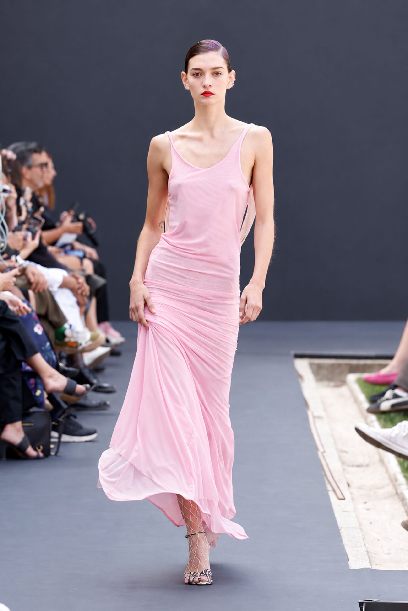 Sheer petal-pink jersey slip dress, ruched low at one hip. White diamond fishnets visible below the swaying hem. Open-toe heels in zebra print with fine ankle ties. Crossing a hotel courtyard just after the pool has been covered.