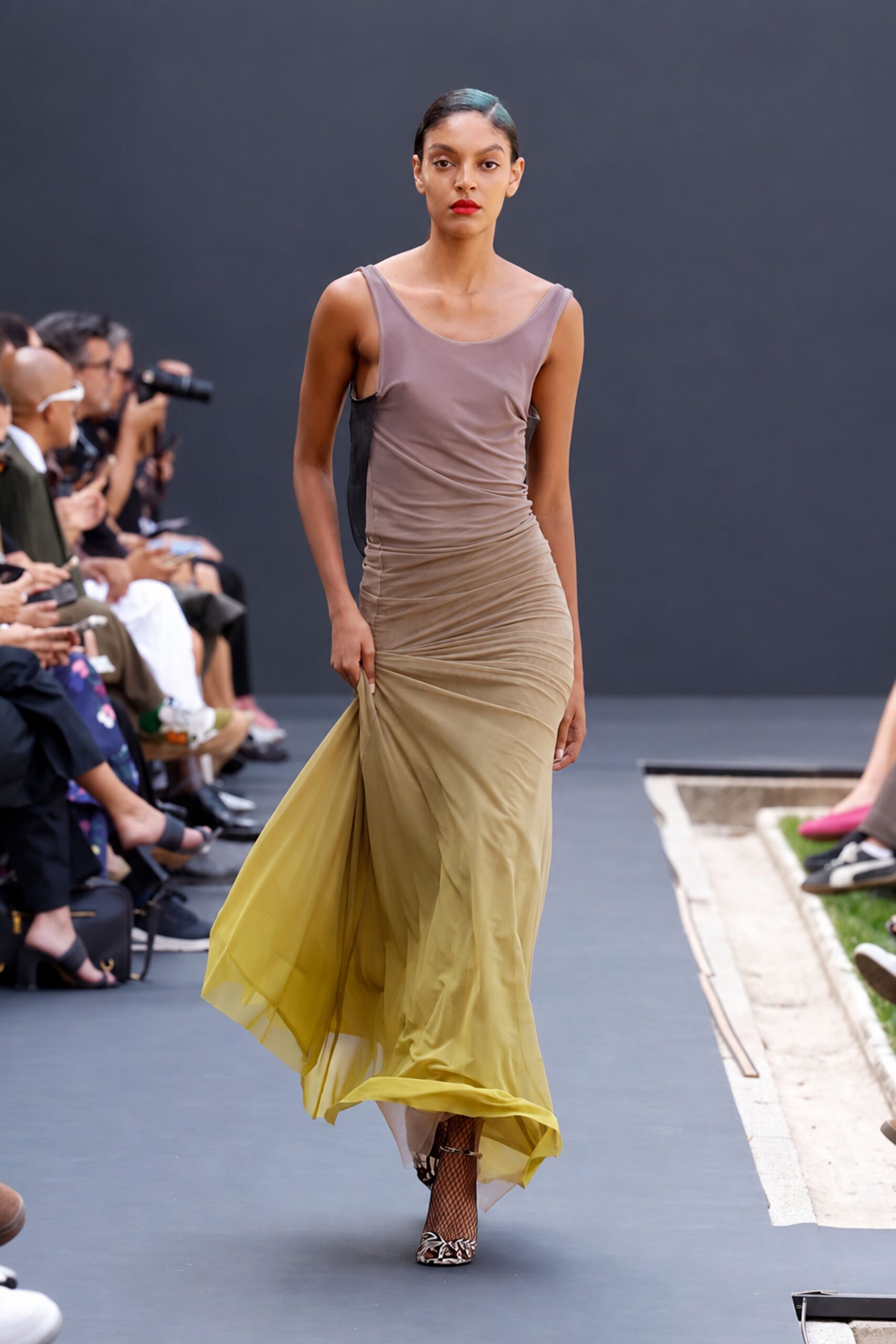 Taupe mesh dress ruched through the hips, fading to chartreuse chiffon. Black fishnet tights under the moving hem. Pointed pumps in zebra pattern, toes peeking past the net. Crossing a museum courtyard just before the storm finally breaks.