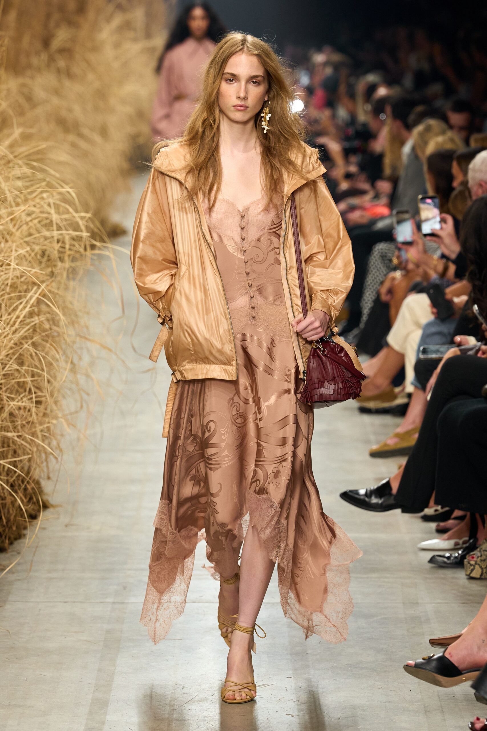 Champagne nylon hooded jacket, softly crinkled with dangling side tabs. Blush devore slip dress, lace insets and tiny covered buttons. Burgundy mini bag with narrow leather fringe. Tan lace-up sandals winding once around the ankle. Single shell-drop earring. Walking back from a beach wedding after the light has gone peach.