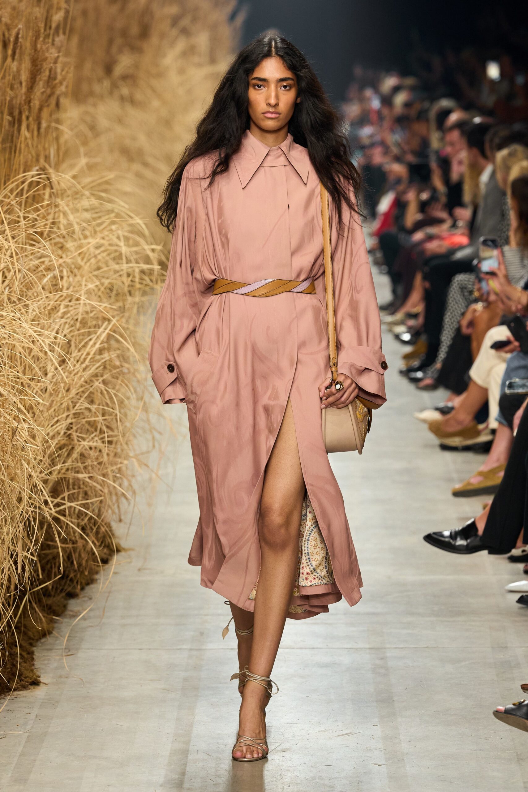Blush satin shirt dress with a wide pointed collar and wrapped front slit. Braided belt in camel and pale lilac crossing the waist. Small beige saddle bag on a tan strap, tucked under the arm. Barely there lace-up sandals winding twice at the ankle. Walking back from a vineyard lunch, dust lifting off the path.