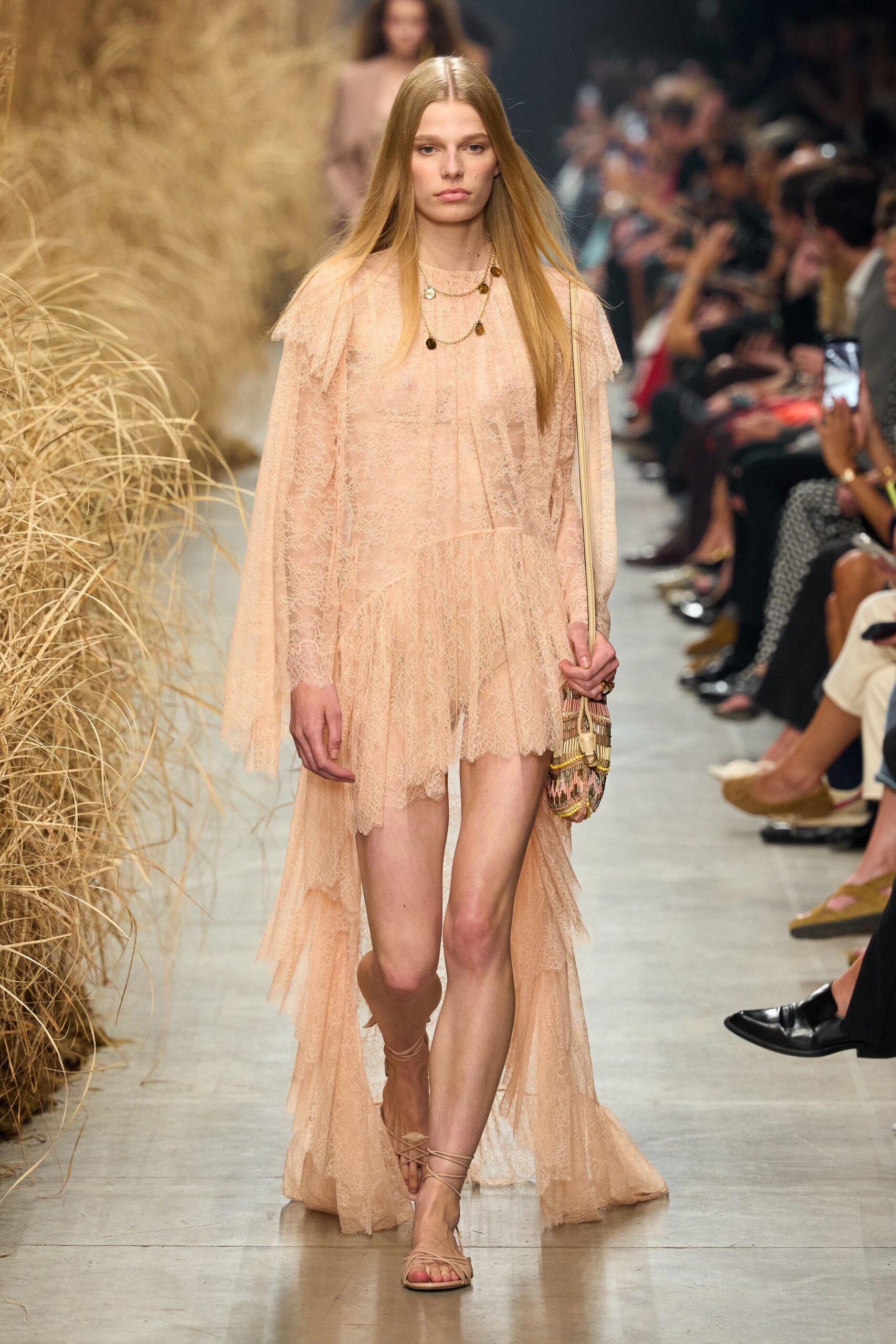 Blush lace dress with cape sleeves and a long trailing back hem. Fine gold chains dotted with small uneven coin charms. Beaded pouch in dusty pink zigzags and cream fringe. Nude wrap sandals laced high around the ankle. Walking across a dry lawn after the wedding, shoes already in hand.